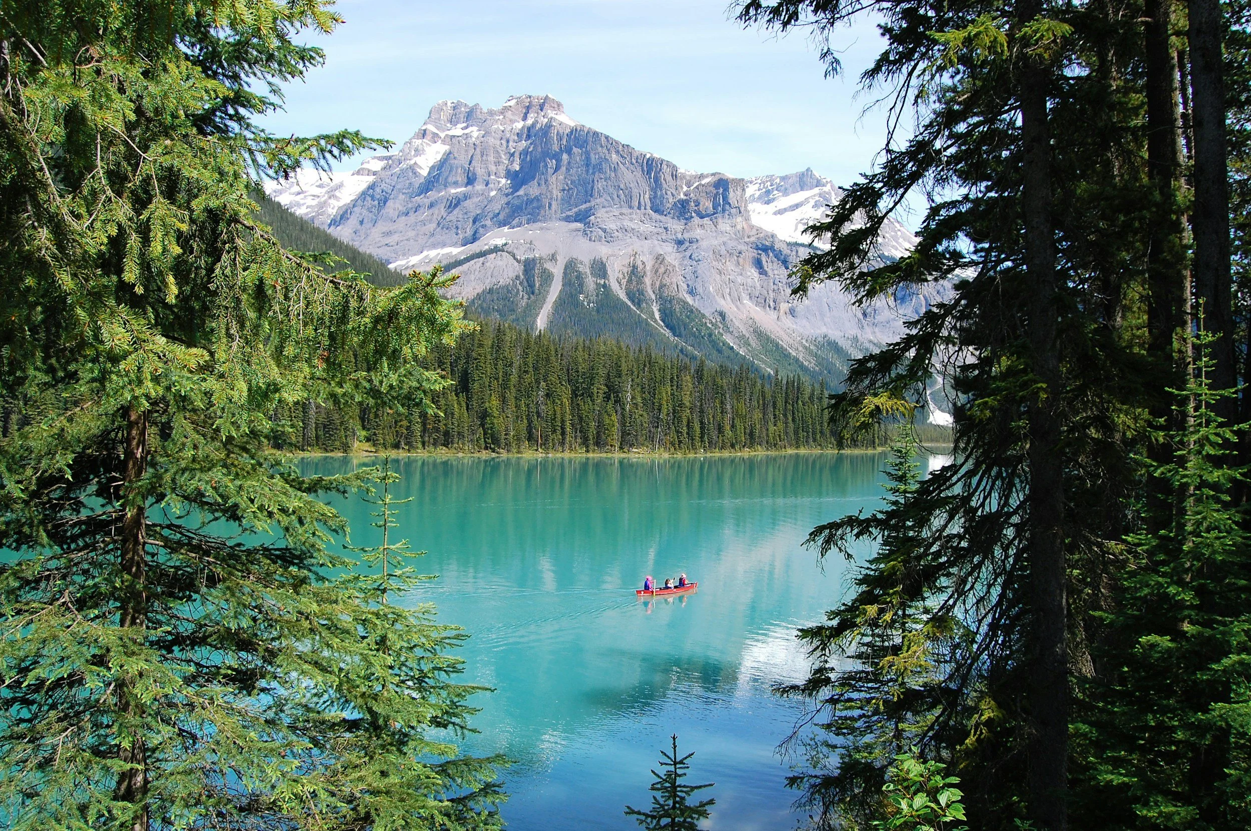 A scenic view of a turquoise lake surrounded by dense evergreen forest with snow-capped mountains in the background. A small red canoe with four people is on the lake.