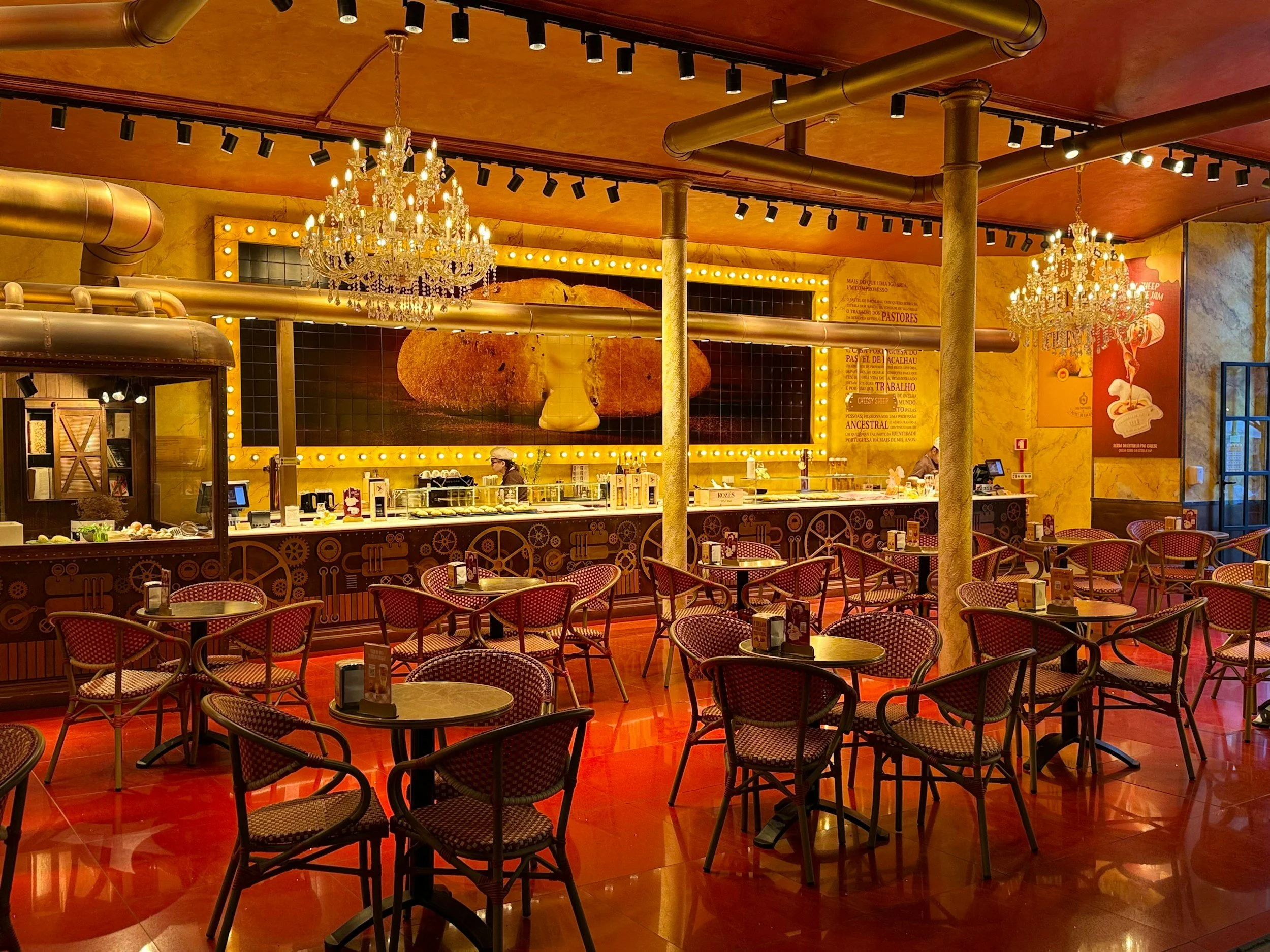 Interior of a restaurant with red patterned chairs, small round tables, a bar counter, chandeliers, and warm yellow walls decorated with large art pieces and signage.