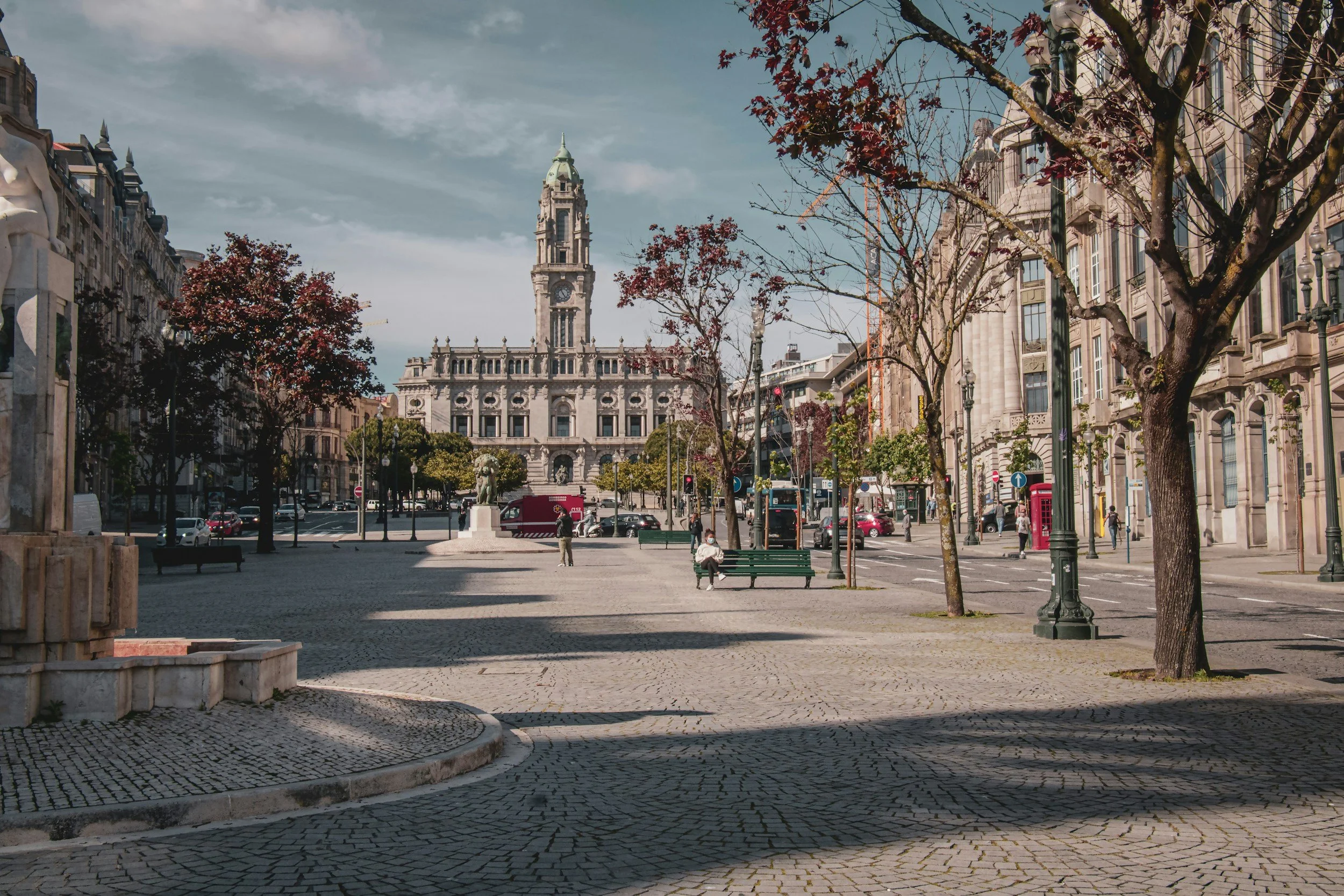 A city square with trees, benches, and a statue in the foreground, and historic buildings with a clock tower in the background, under a partly cloudy sky.