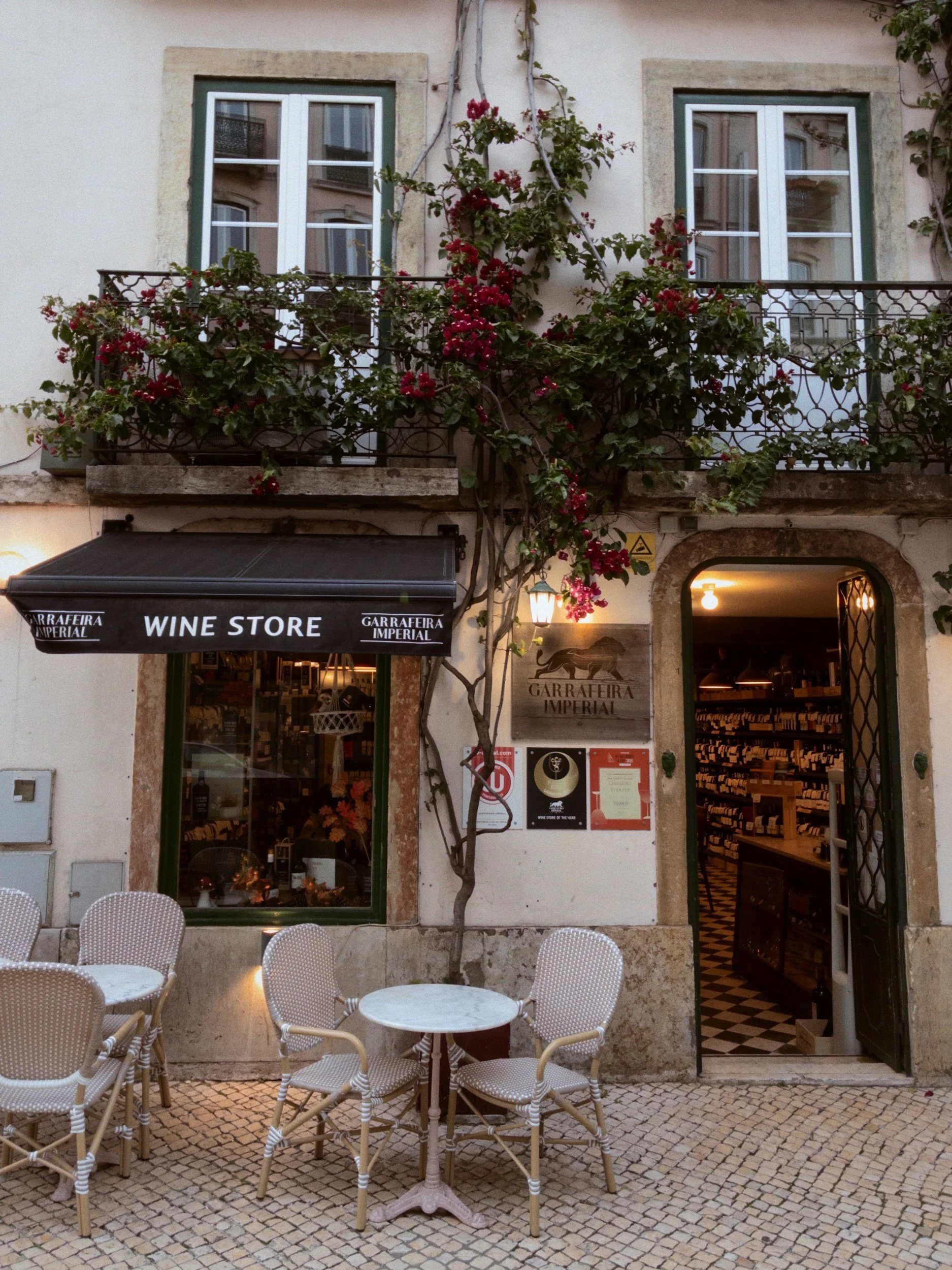 Exterior of a wine store with outdoor seating on cobblestone pavement, surrounded by flowering vines and a balcony above.