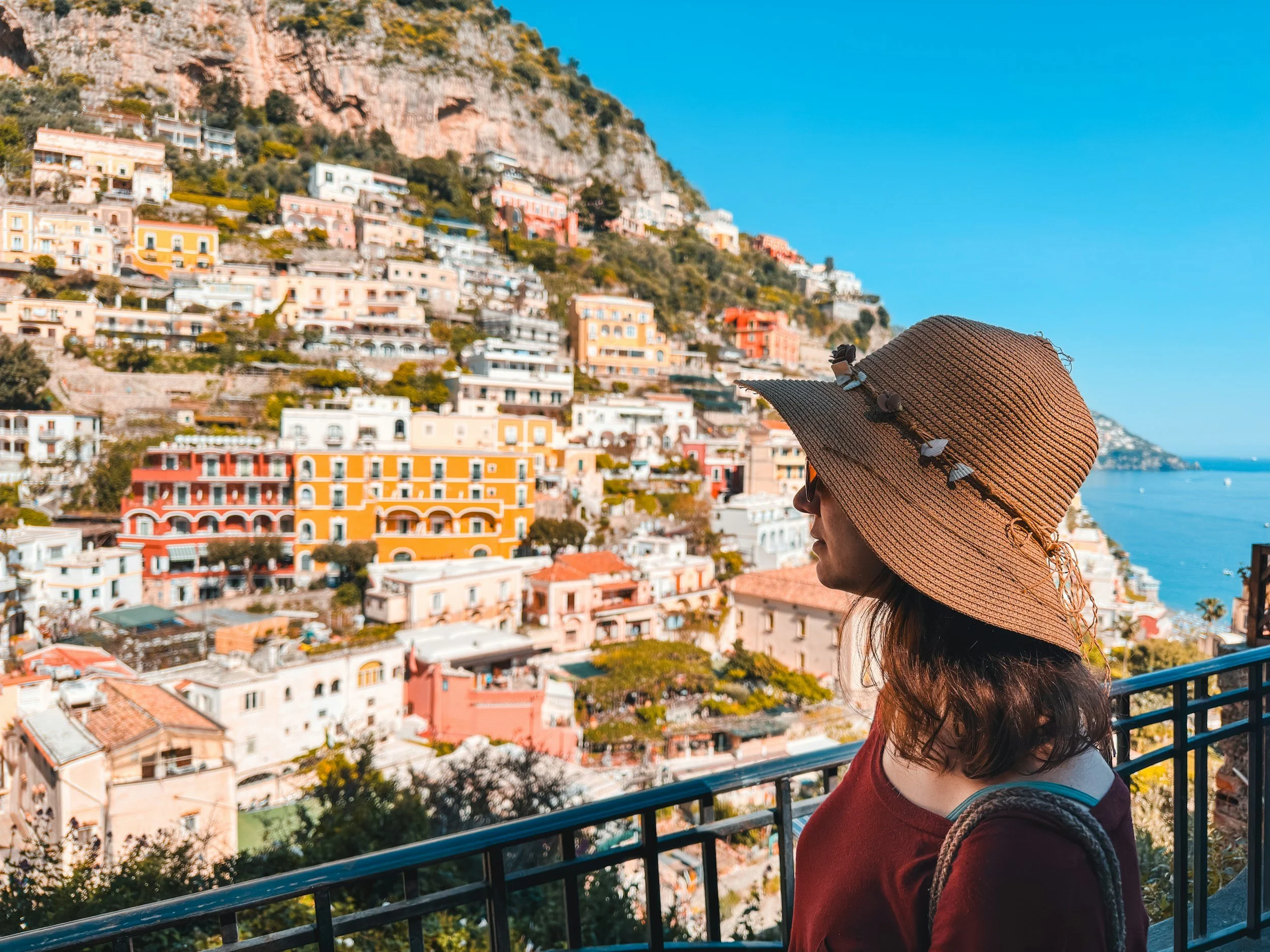 A woman wearing a wide-brimmed hat and sunglasses standing on a balcony overlooking a colorful hillside town by the sea.