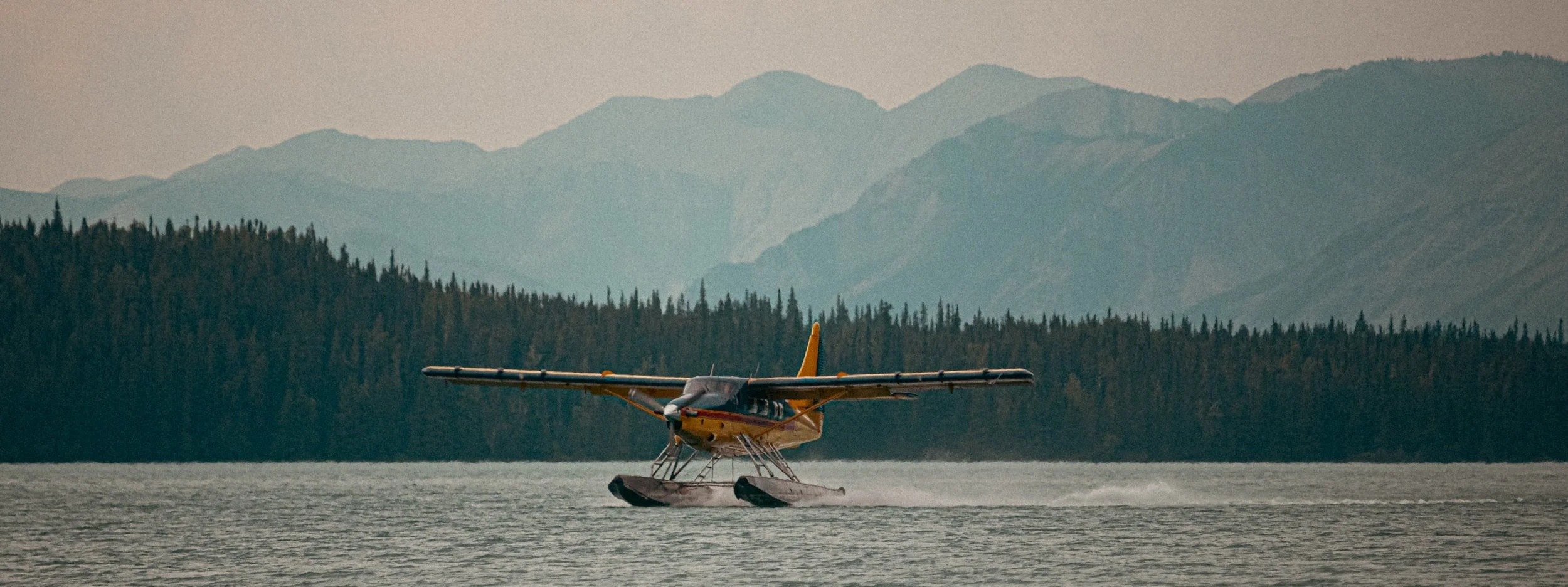 Yellow seaplane flying over a body of water with mountains and a forested shoreline in the background during daytime.