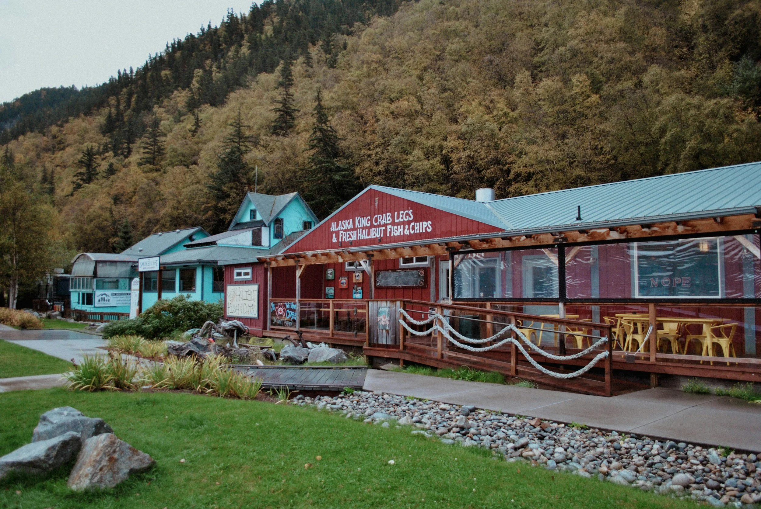 Colorful buildings with turquoise and red exteriors, large windows, and outdoor seating area with yellow chairs, set against a backdrop of forested mountains.