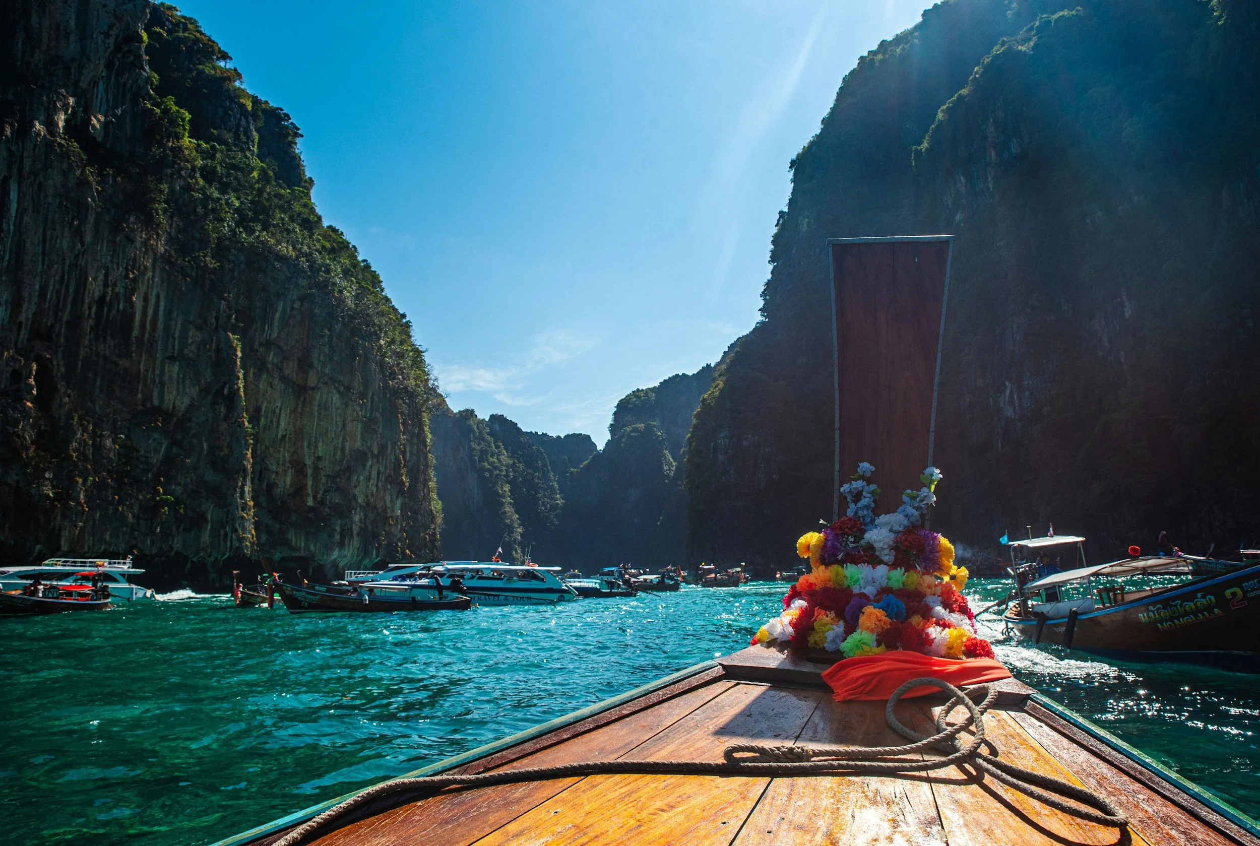 Boats and a floating shrine decorated with colorful flowers on a river surrounded by green cliffs under a clear blue sky.