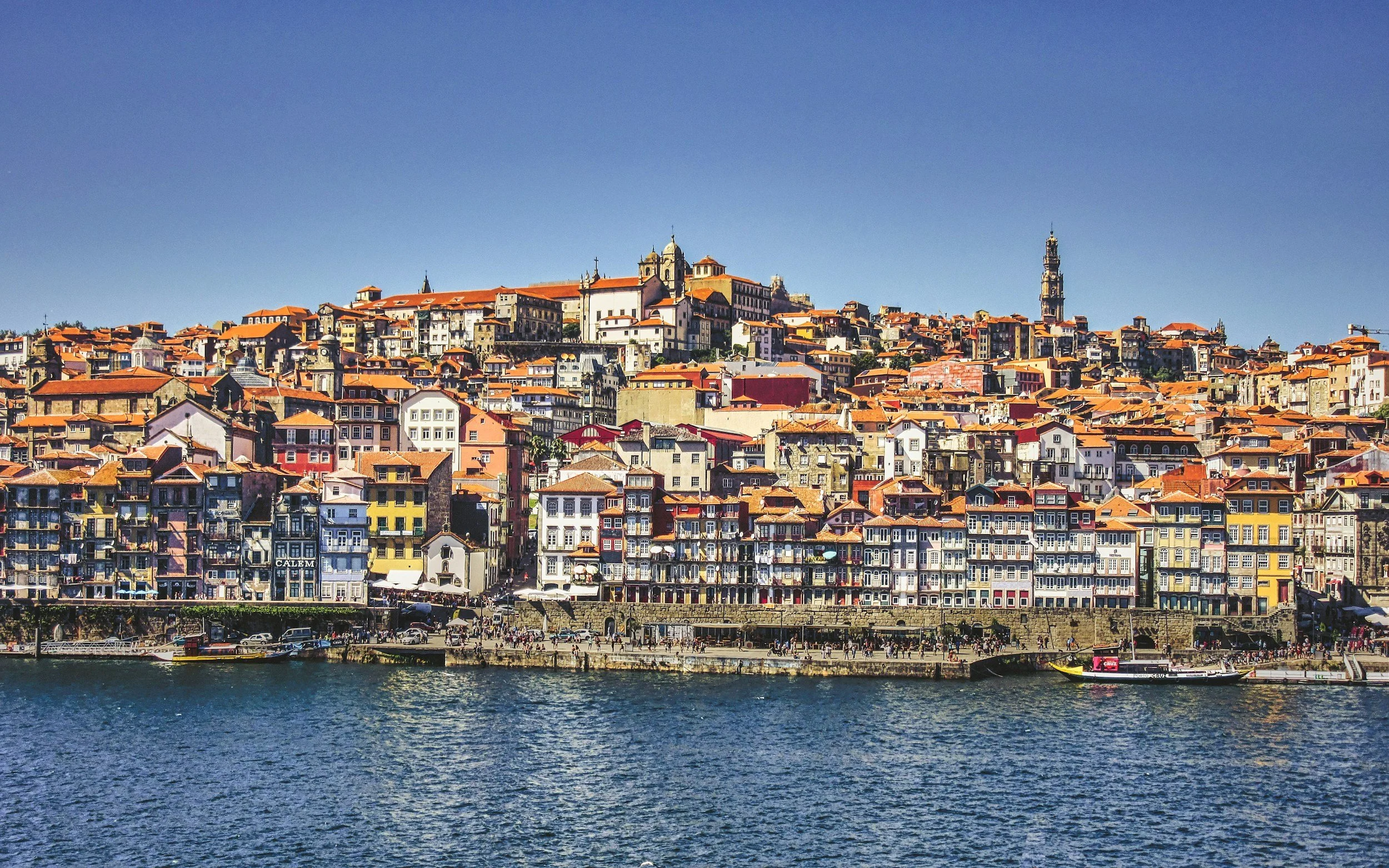 A panoramic view of a city built on a hill with colorful buildings topped with red-tiled roofs, seen from across a river with boats docked along the shore.