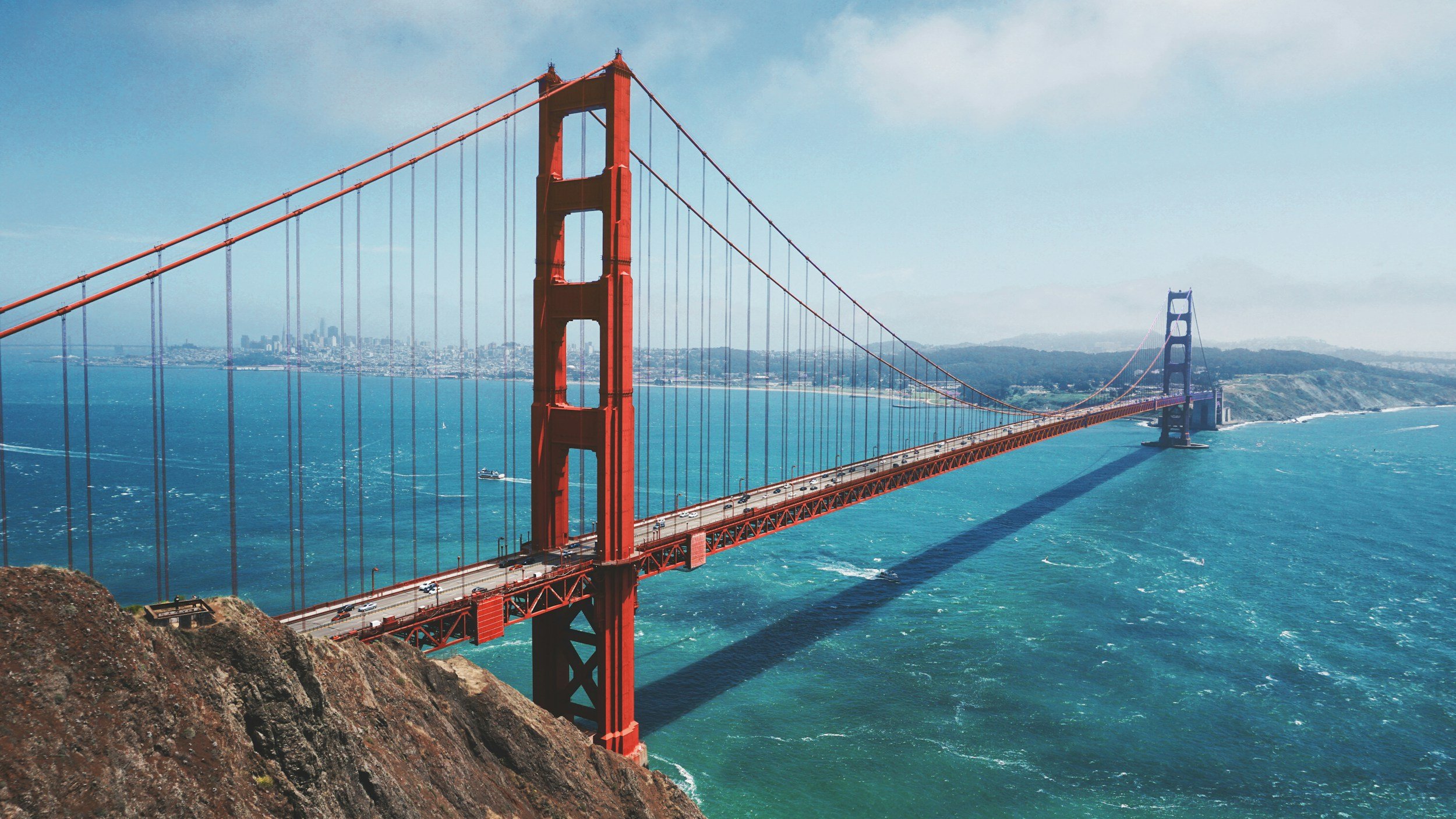 The Golden Gate Bridge in San Francisco, California, seen from above with the city and hills in the background. The bridge is painted red and spans across the blue water of the bay, with cars traveling on it.