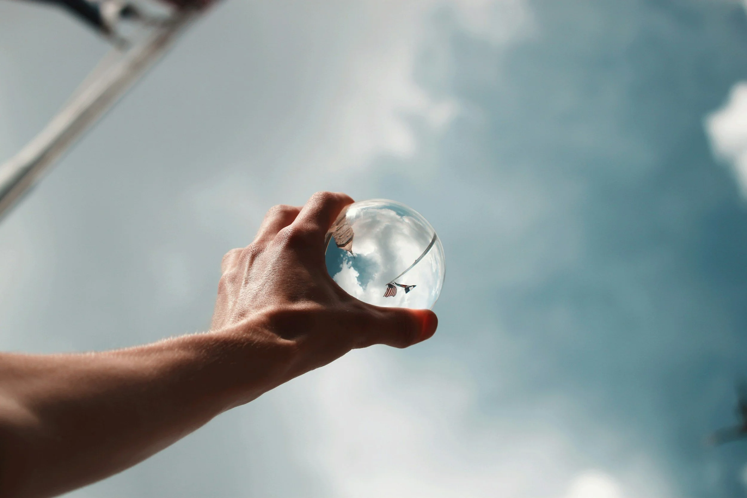 Close-up of a person's hand holding a transparent glass with a reflection of clouds and American flags, against a partly cloudy sky.