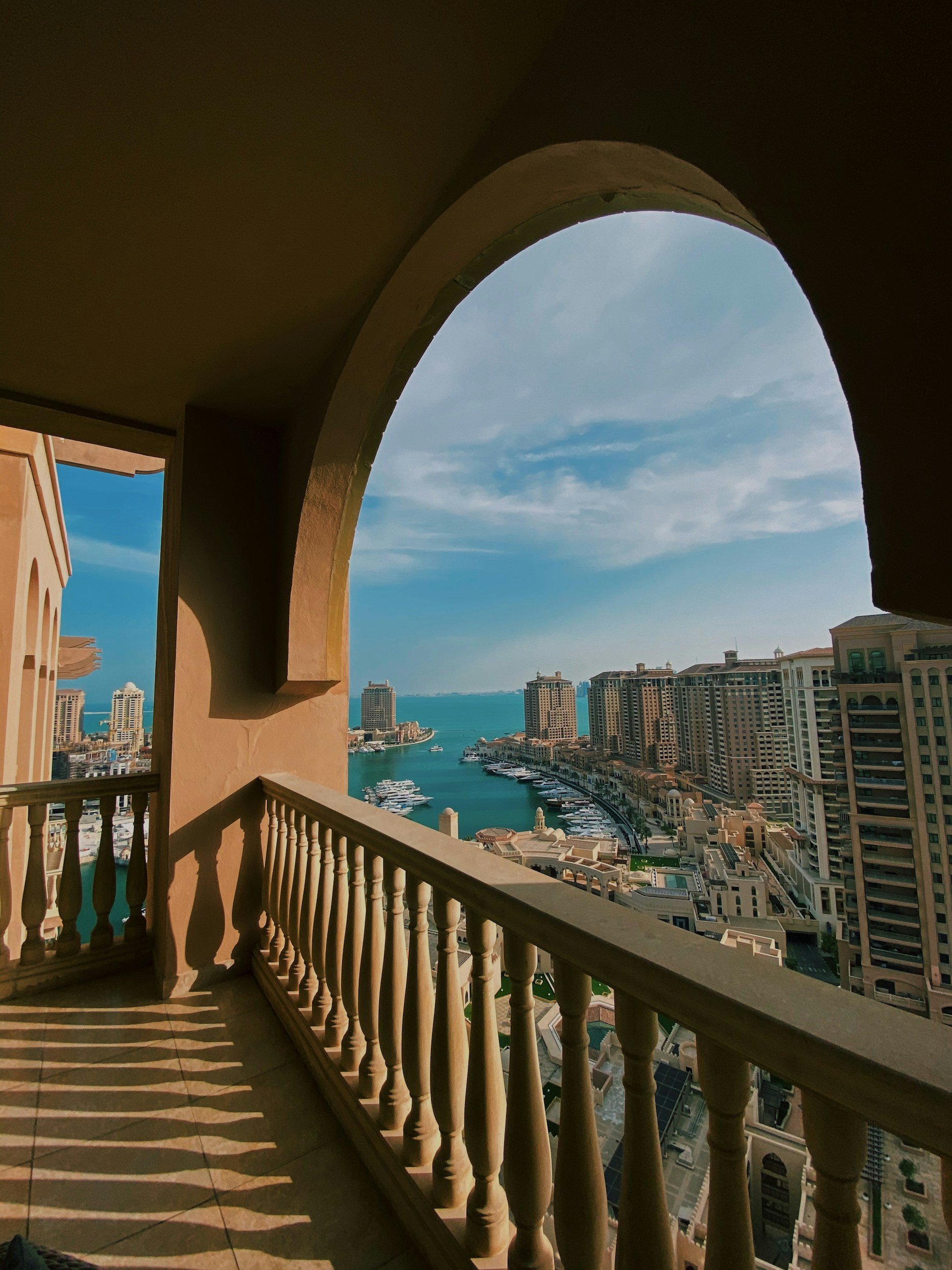 A view from a balcony over a cityscape featuring tall buildings and a waterway, with a blue sky and some clouds, through an arched opening and balustrade.
