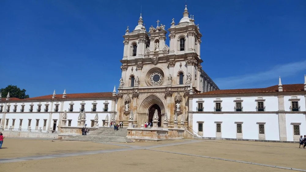 A historic cathedral with ornate architecture and a large central entrance, surrounded by a courtyard with a few people walking. The building features twin bell towers and a cross on top, set against a clear blue sky.