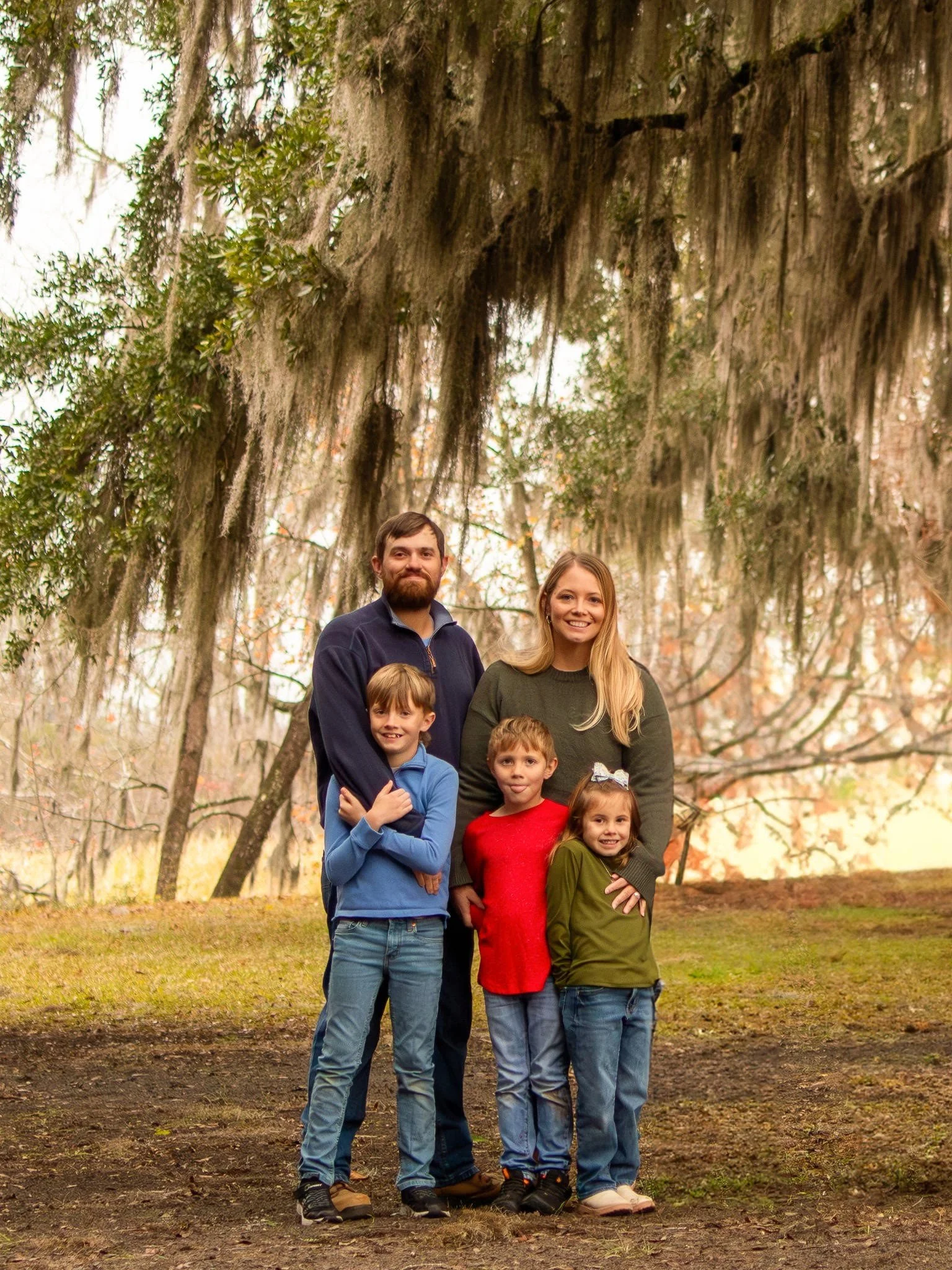 A family of six posing outdoors in front of tall trees with hanging moss, under a large, overhanging branch.