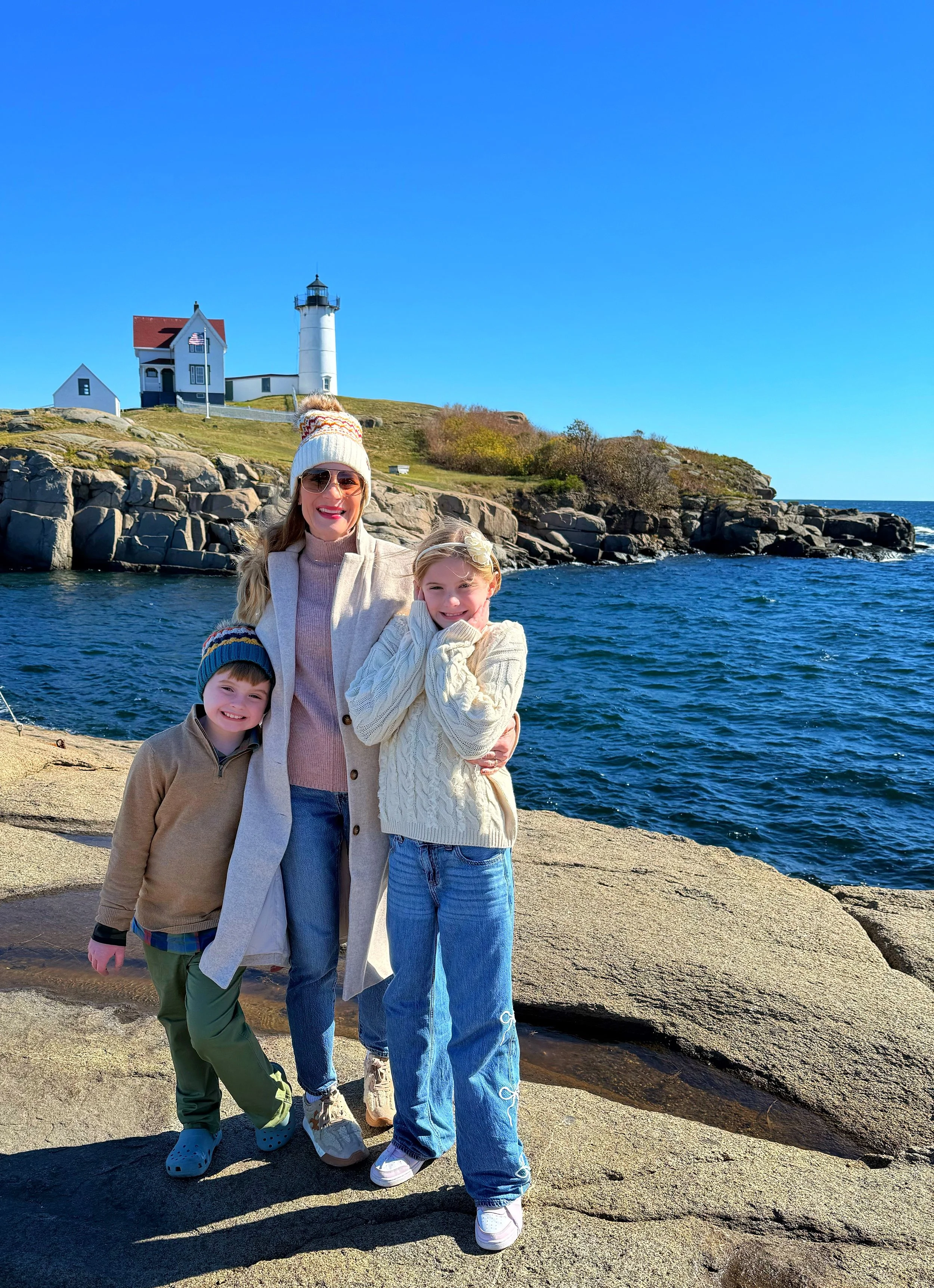 A woman with three children standing on rocks near the water with a lighthouse and houses in the background on a sunny day.