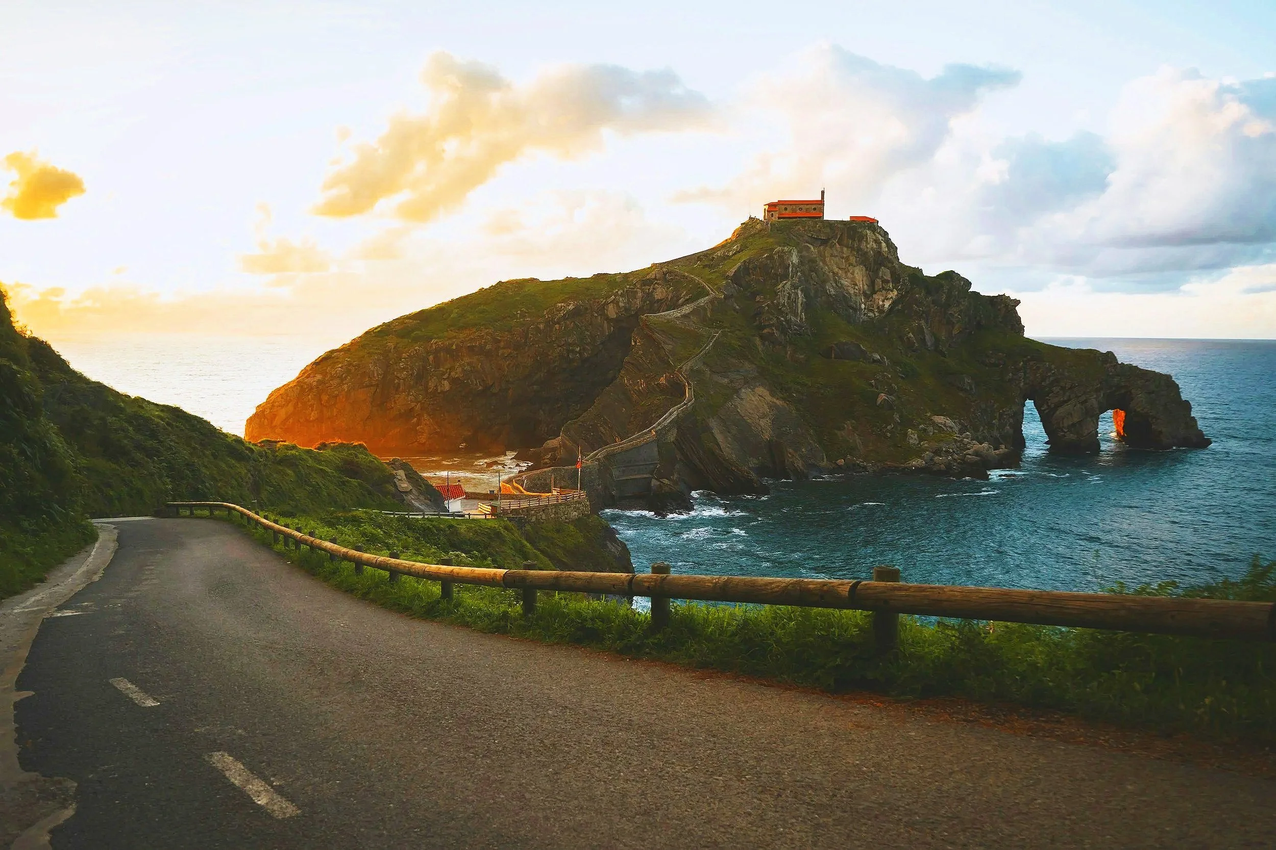 A coastal road curves around green cliffs leading to a large rocky island with arches and a building at the top, illuminated by sunlight.