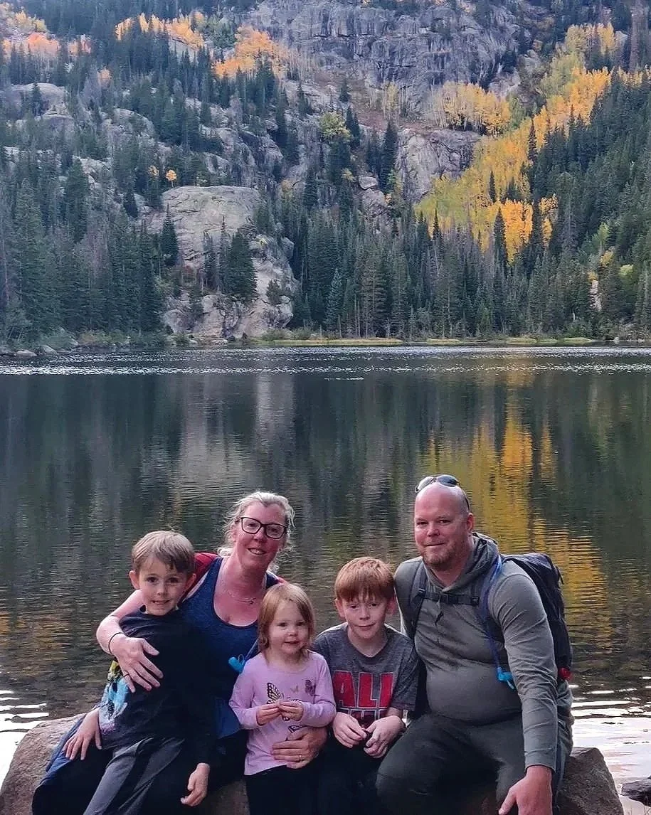 A family of five sitting on rocks by a lake with a mountain and forest in the background, autumn colors in the trees.