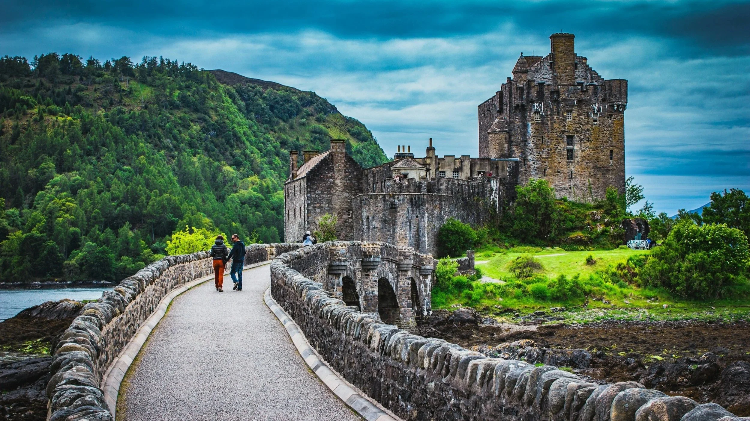 A stone historic castle on a hill with lush green trees, connected by a narrow stone bridge with a railing over water, with a cloudy sky overhead.