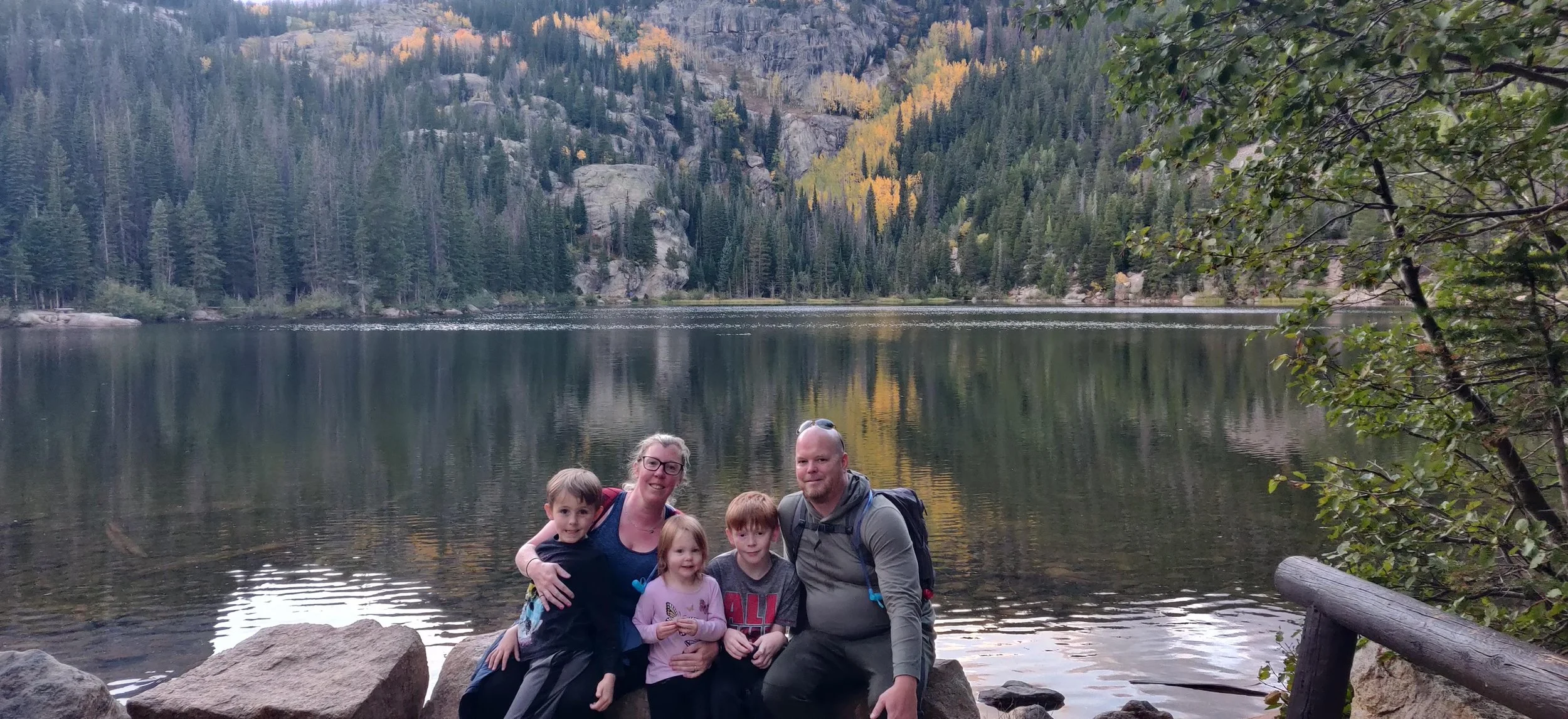 Family of five, including two parents and three children, sitting on rocks near a lake with a forested mountain landscape in the background.