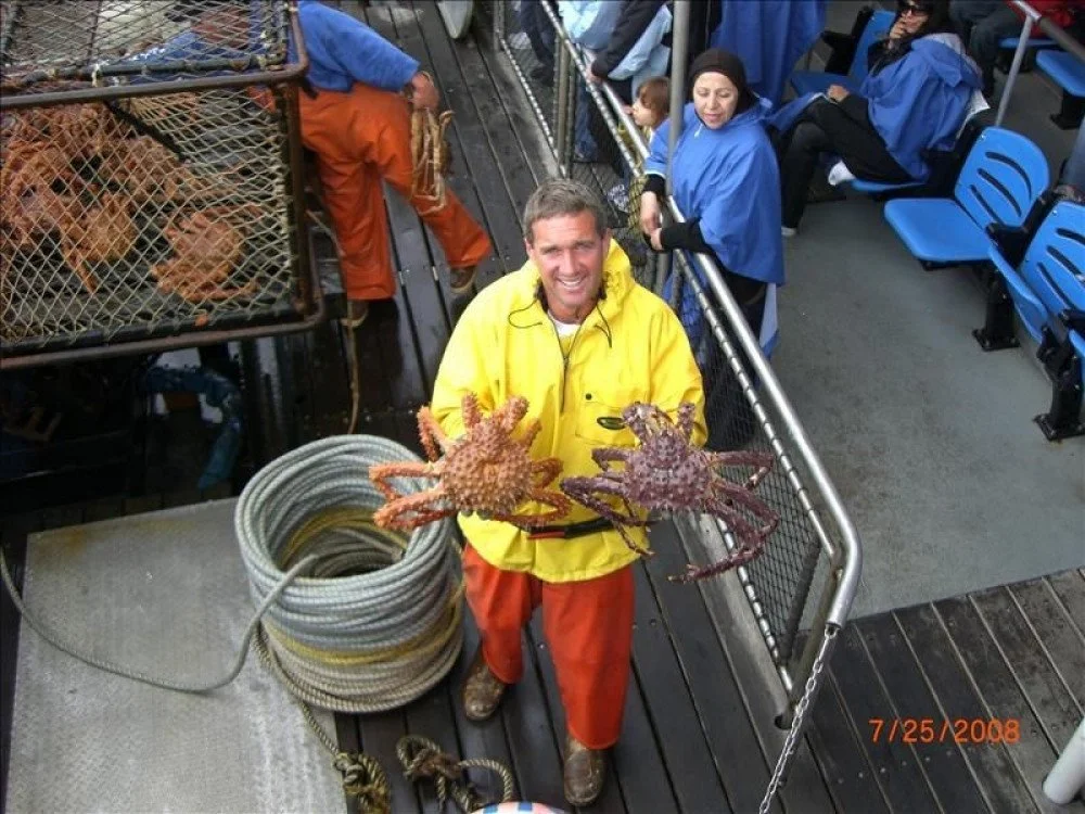A man in a yellow raincoat and orange pants holding two large octopuses on a ship deck, with other people in rain jackets in the background and a cage with lobsters or crabs nearby.