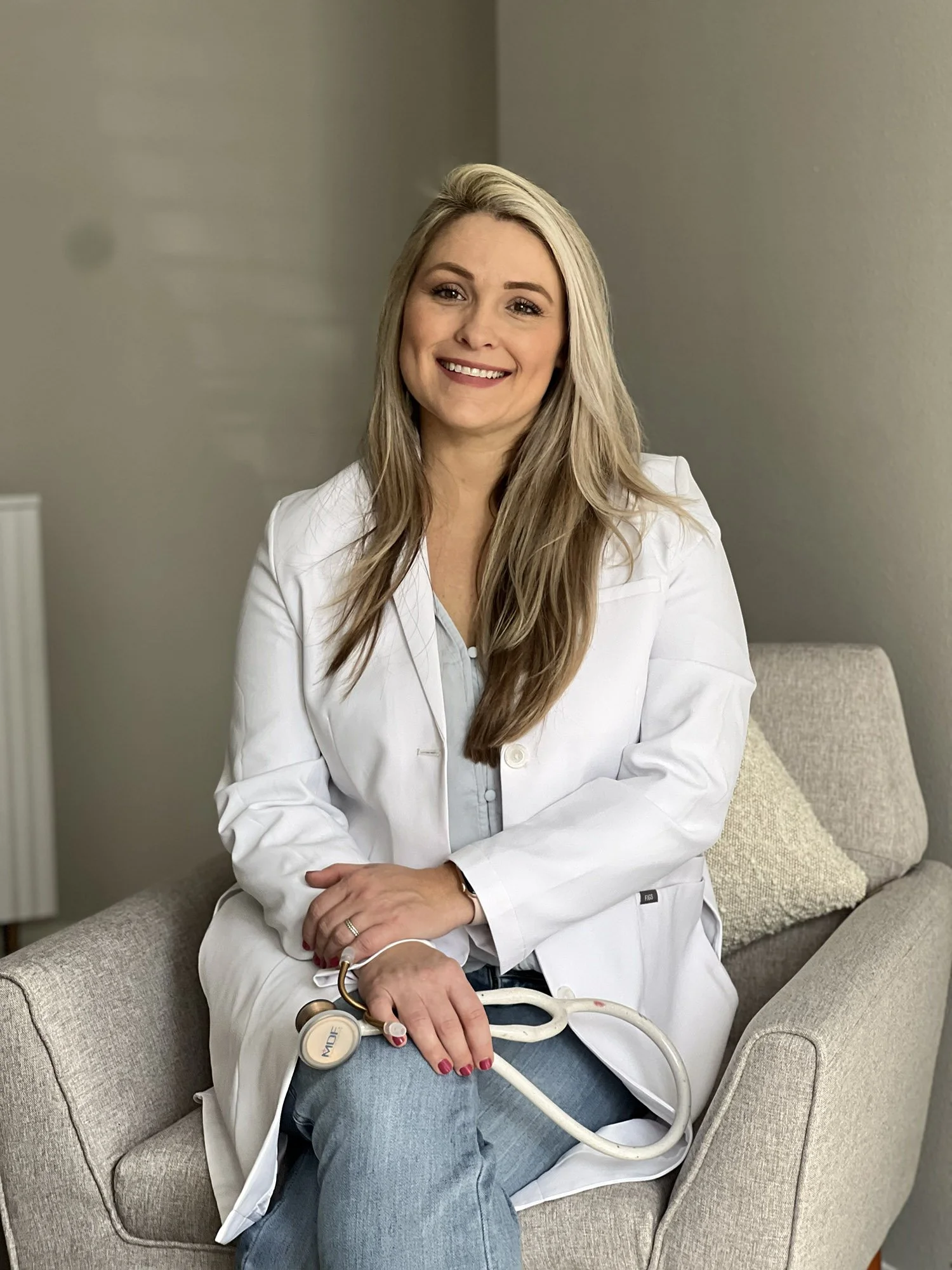 A smiling female doctor or healthcare professional with long blonde hair, wearing a white lab coat, sitting on a beige armchair with a stethoscope resting on her lap, in a neutral-colored room.