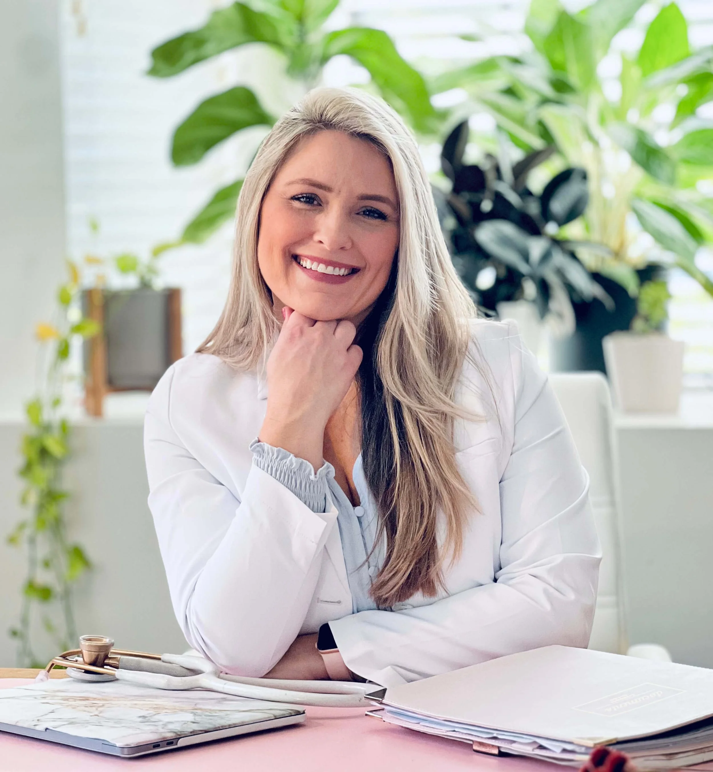 A smiling female doctor with long blonde hair, wearing a white coat, sitting at a desk with medical documents and a stethoscope, in a bright, plant-filled office.
