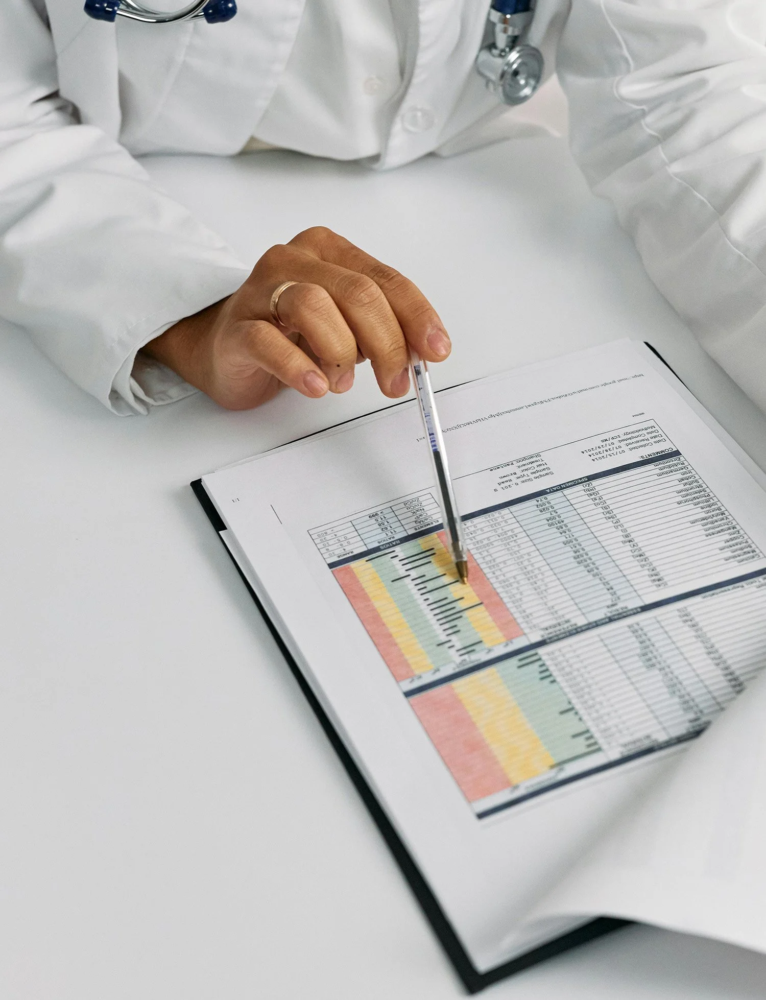 A medical professional in a white coat points to a colorful chart or table on a clipboard, with a stethoscope draped around their neck.
