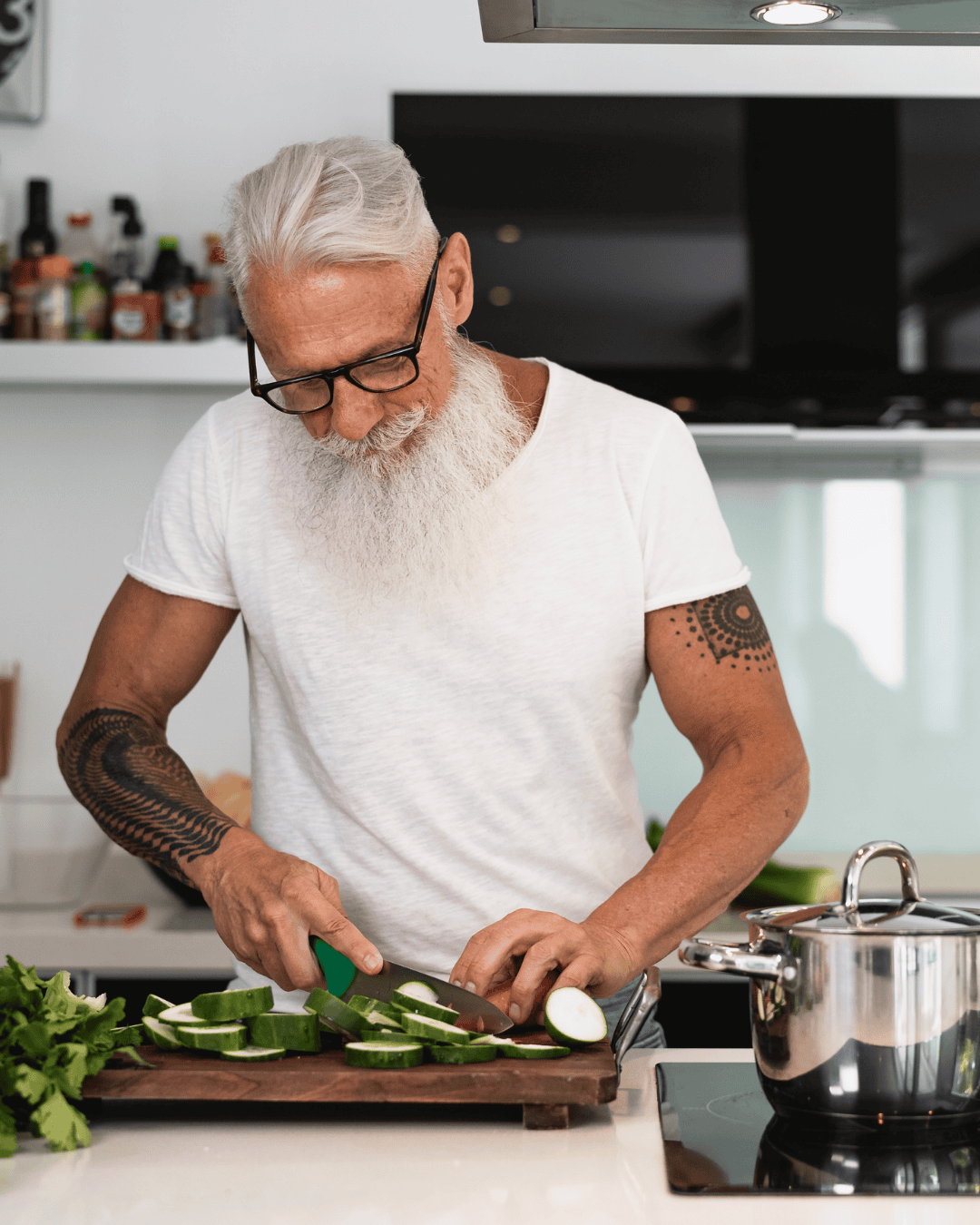 An elderly man with a white beard and glasses is chopping cucumbers on a kitchen cutting board. He is wearing a white t-shirt and has tattoos on his arms. There are fresh greens and a pot on the stove nearby.