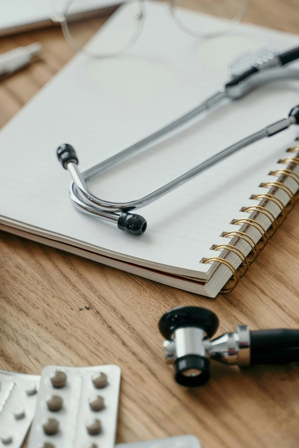 A stethoscope and a pair of glasses resting on an open spiral notebook, with blister packs of pills nearby on a wooden surface.