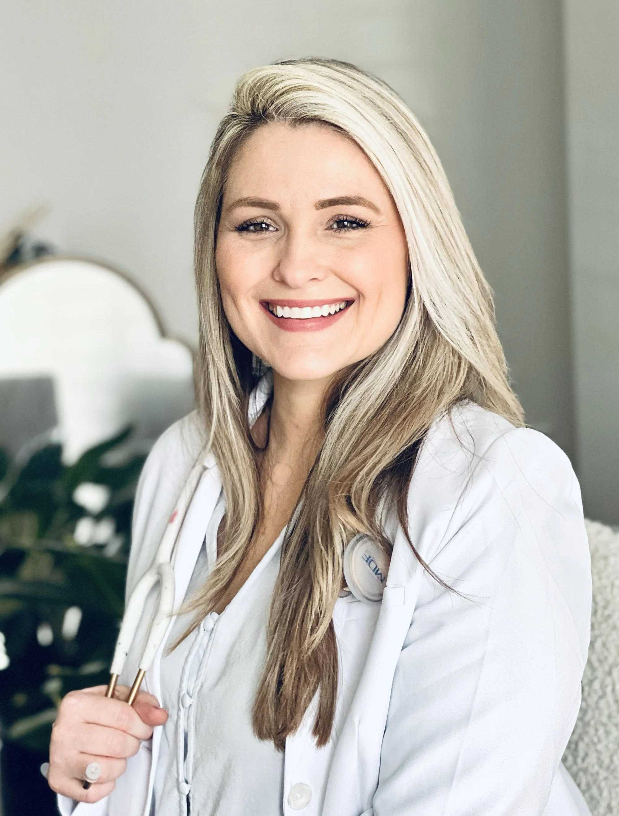 A smiling female doctor with long blonde hair, wearing a white lab coat and stethoscope, in a bright room.