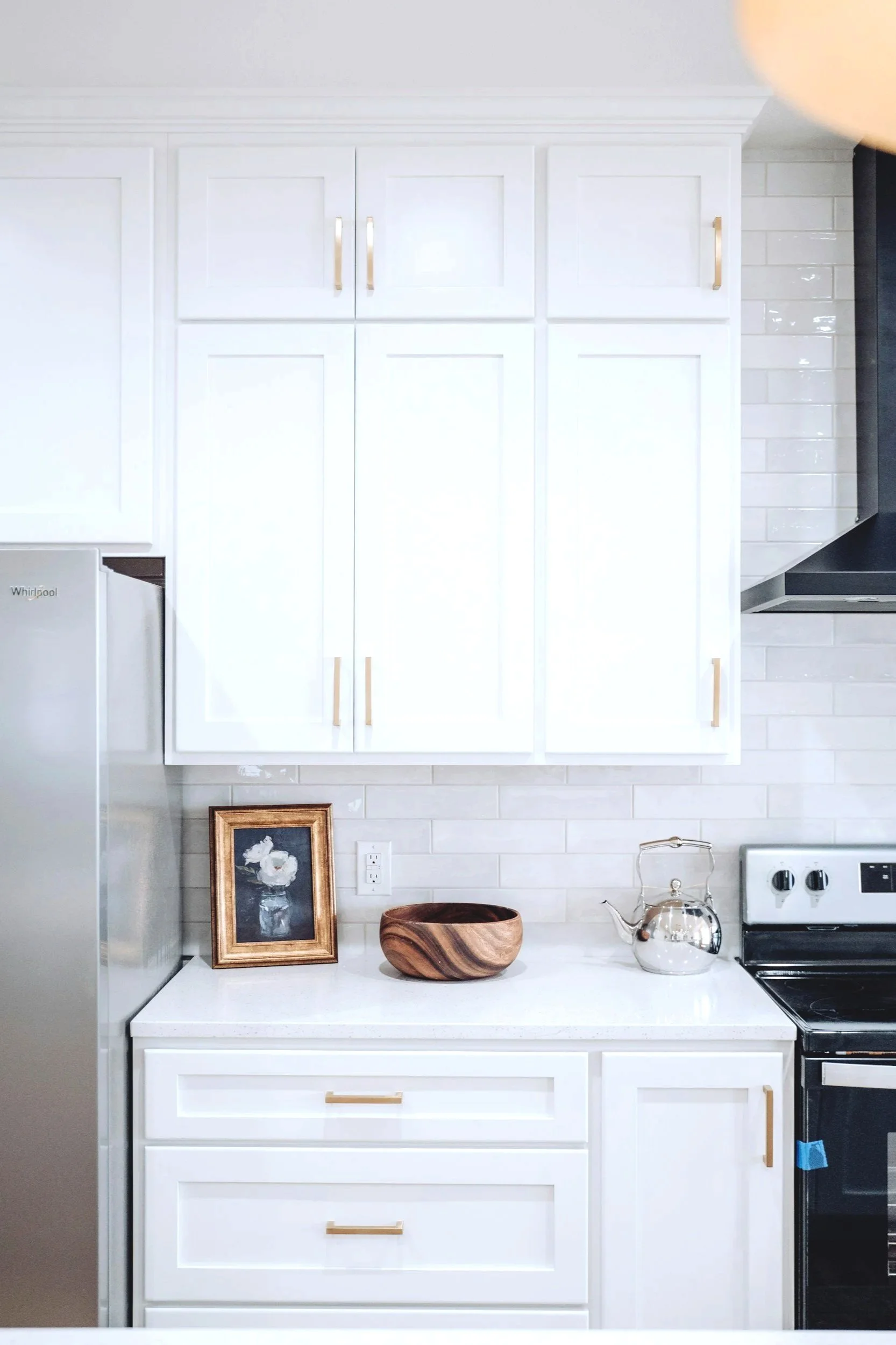 White kitchen cabinets with gold handles, a small framed picture, a wooden bowl, a silver teapot, and a white oven with black stovetop in a modern kitchen.