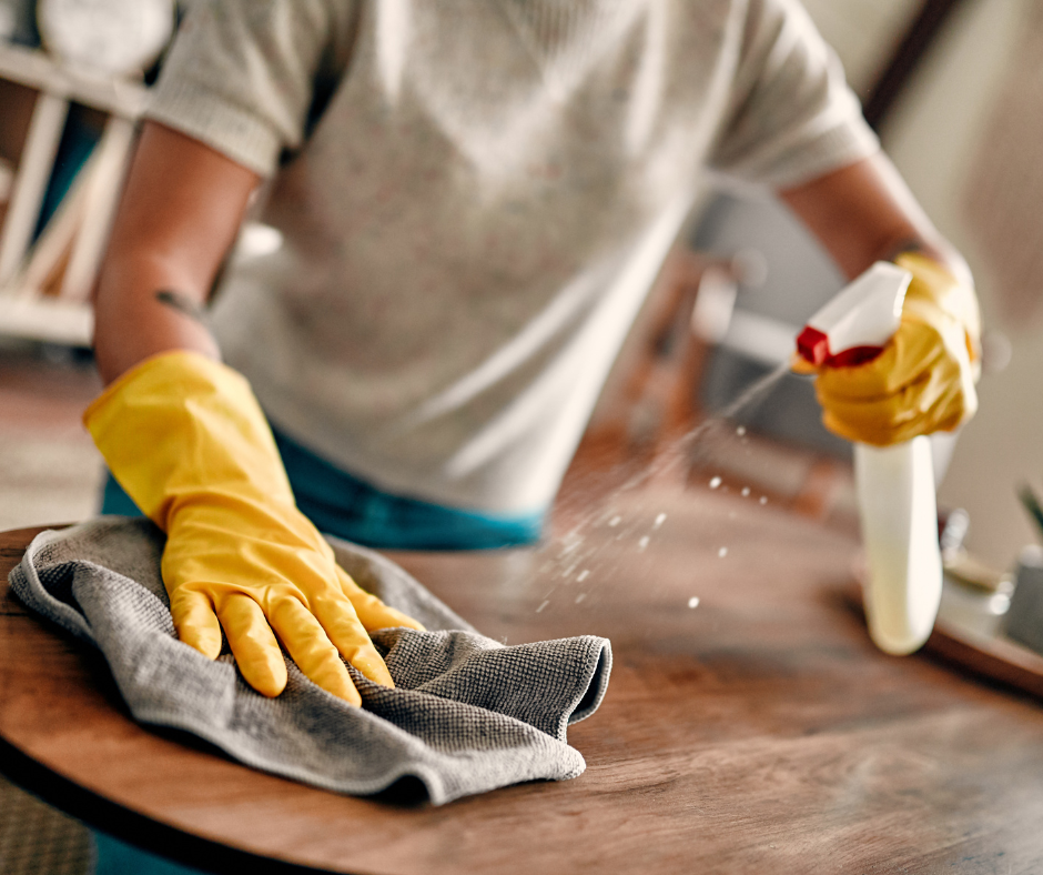 Person wearing yellow rubber gloves cleaning a wooden table with a gray cloth spray bottle.