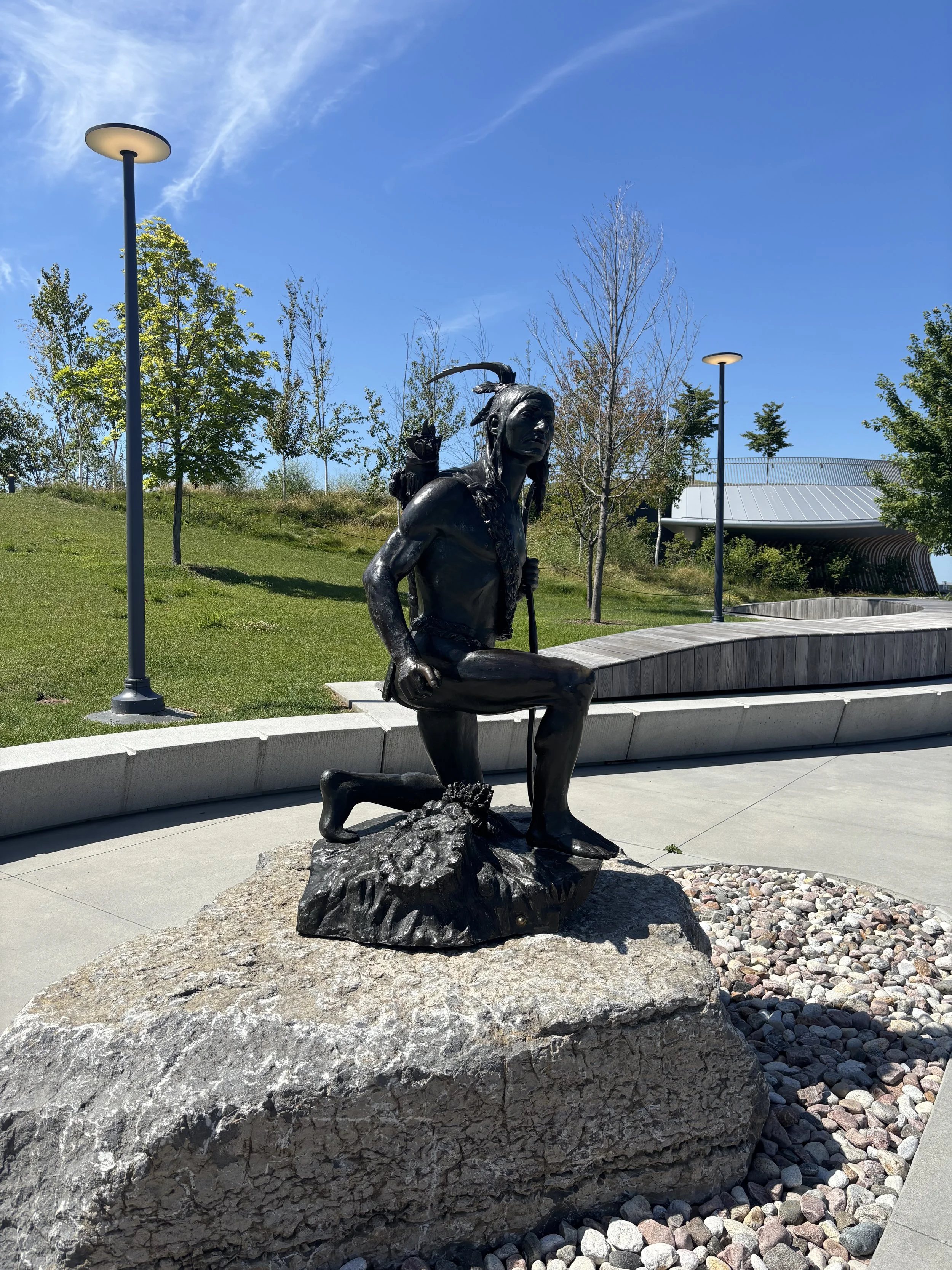 Bronze statue of a Native American man with a feather headdress, kneeling on one knee, with a rifle slung over his shoulder, set on a large stone in a park with trees, benches, and modern street lamps under a blue sky.