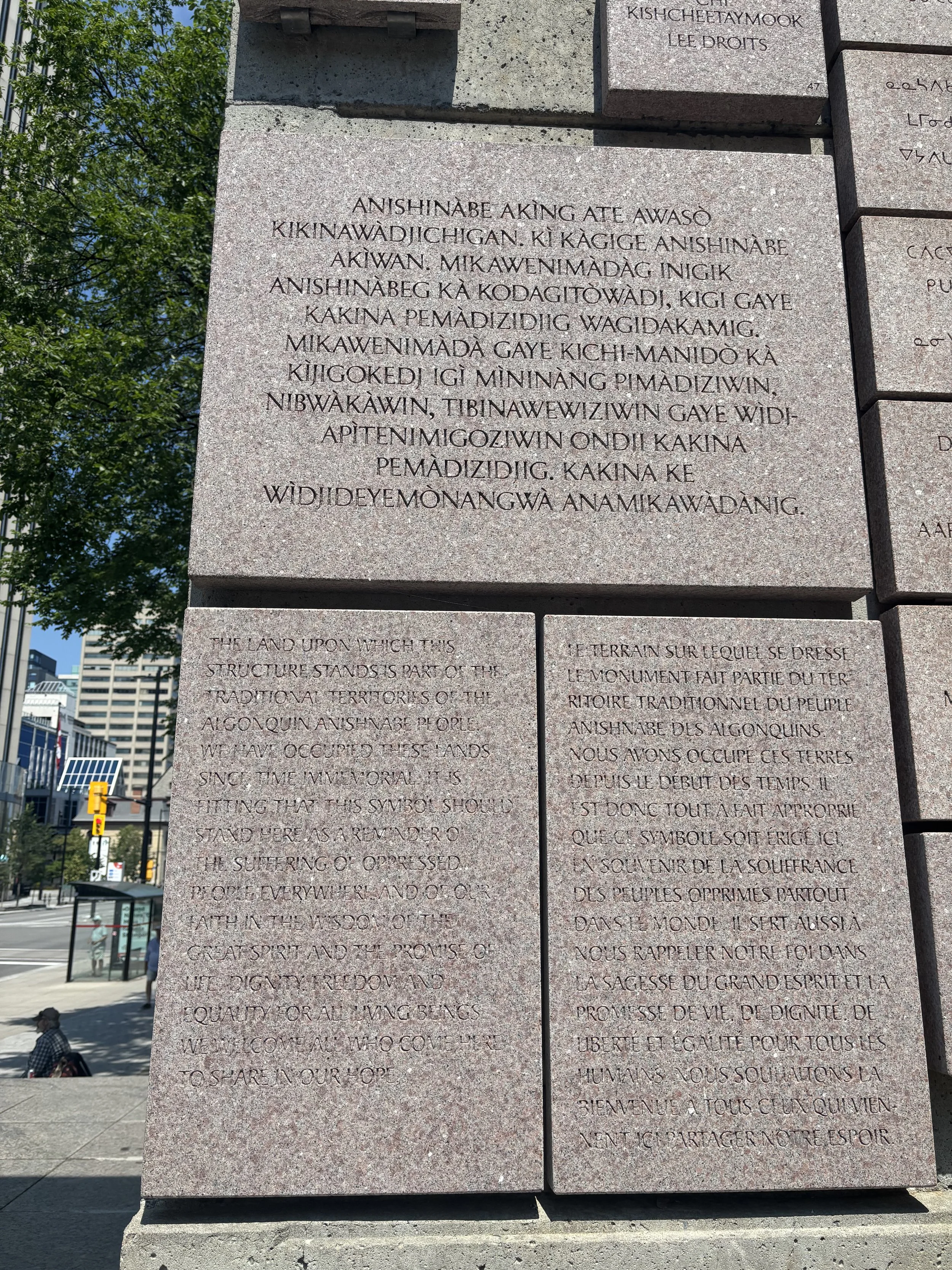 Text on a stone monument with inscriptions in English and another language, situated outdoors in an urban area with buildings, trees, and a person sitting on a bench nearby.