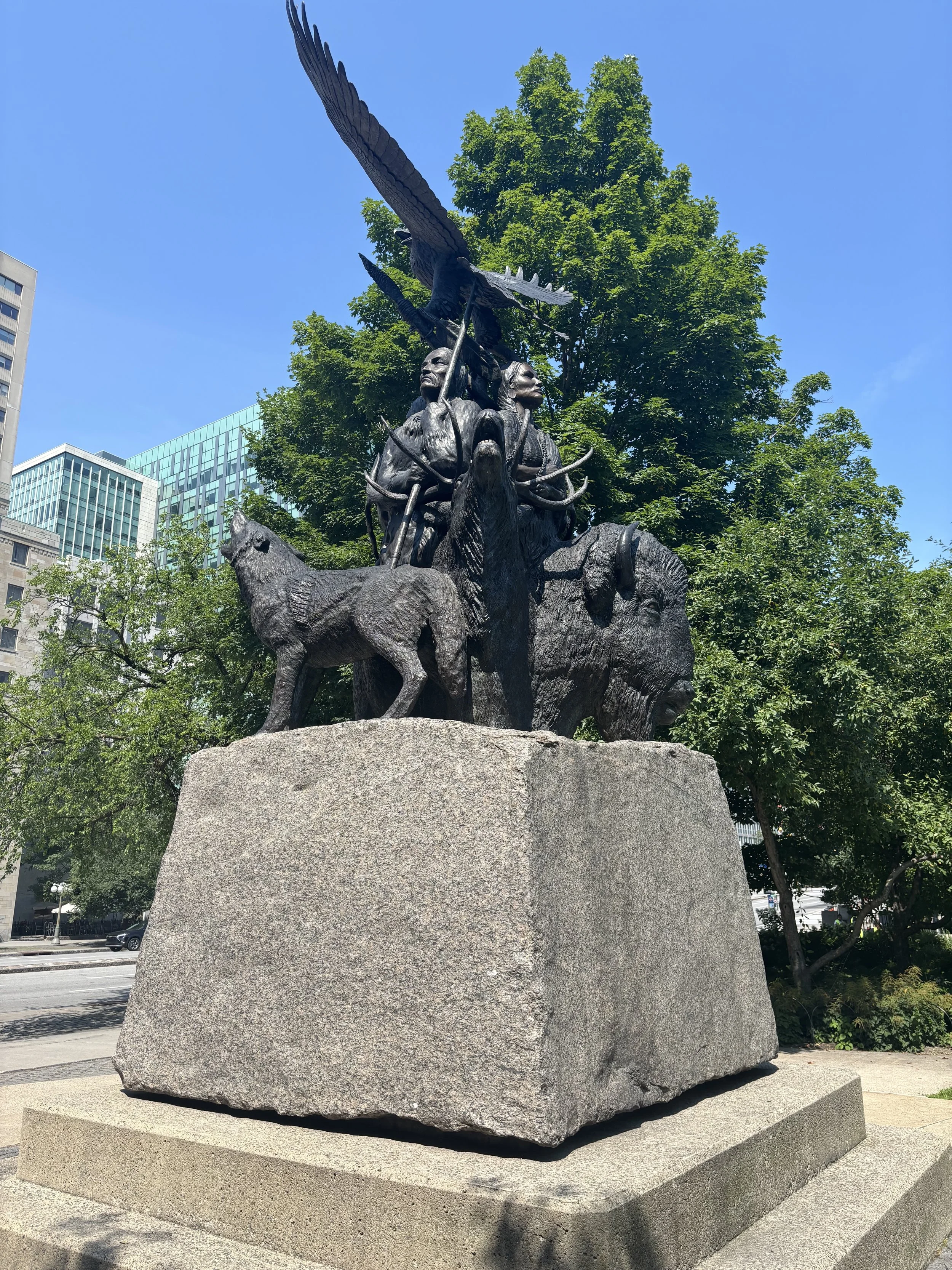 Bronze monument depicting Indigenous people with animals and an eagle, set on a large stone base in a city park with modern buildings and greenery in the background.
