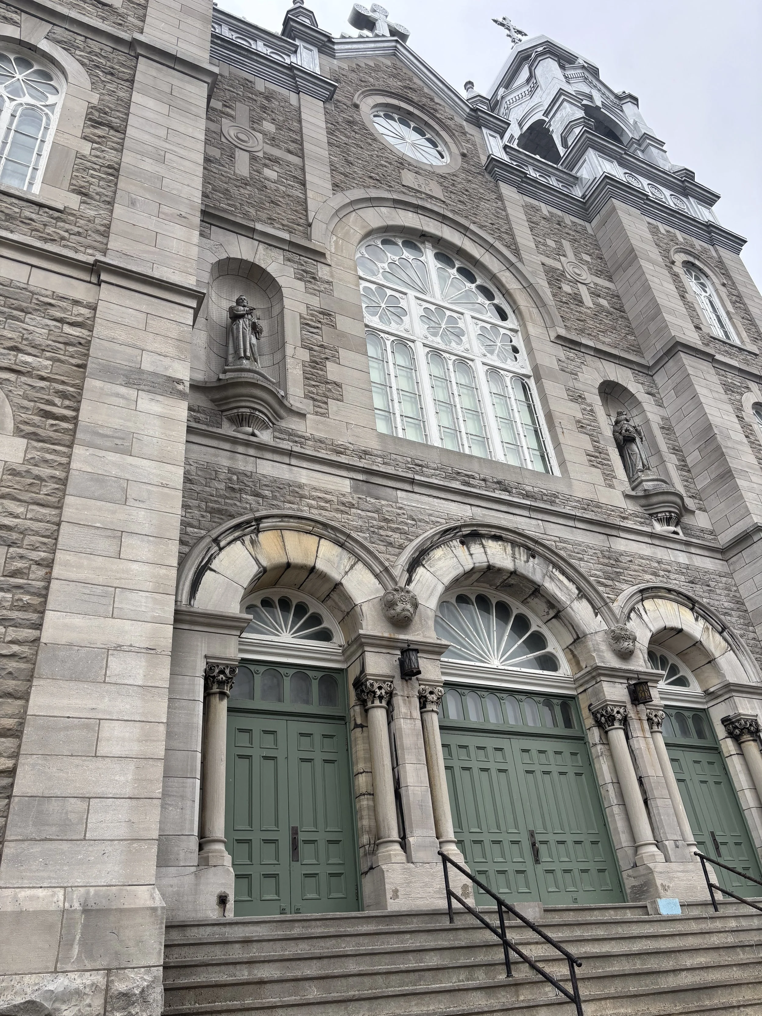 Front view of a stone church with three green doors, arched windows, decorative columns, statues in niches, and a bell tower.