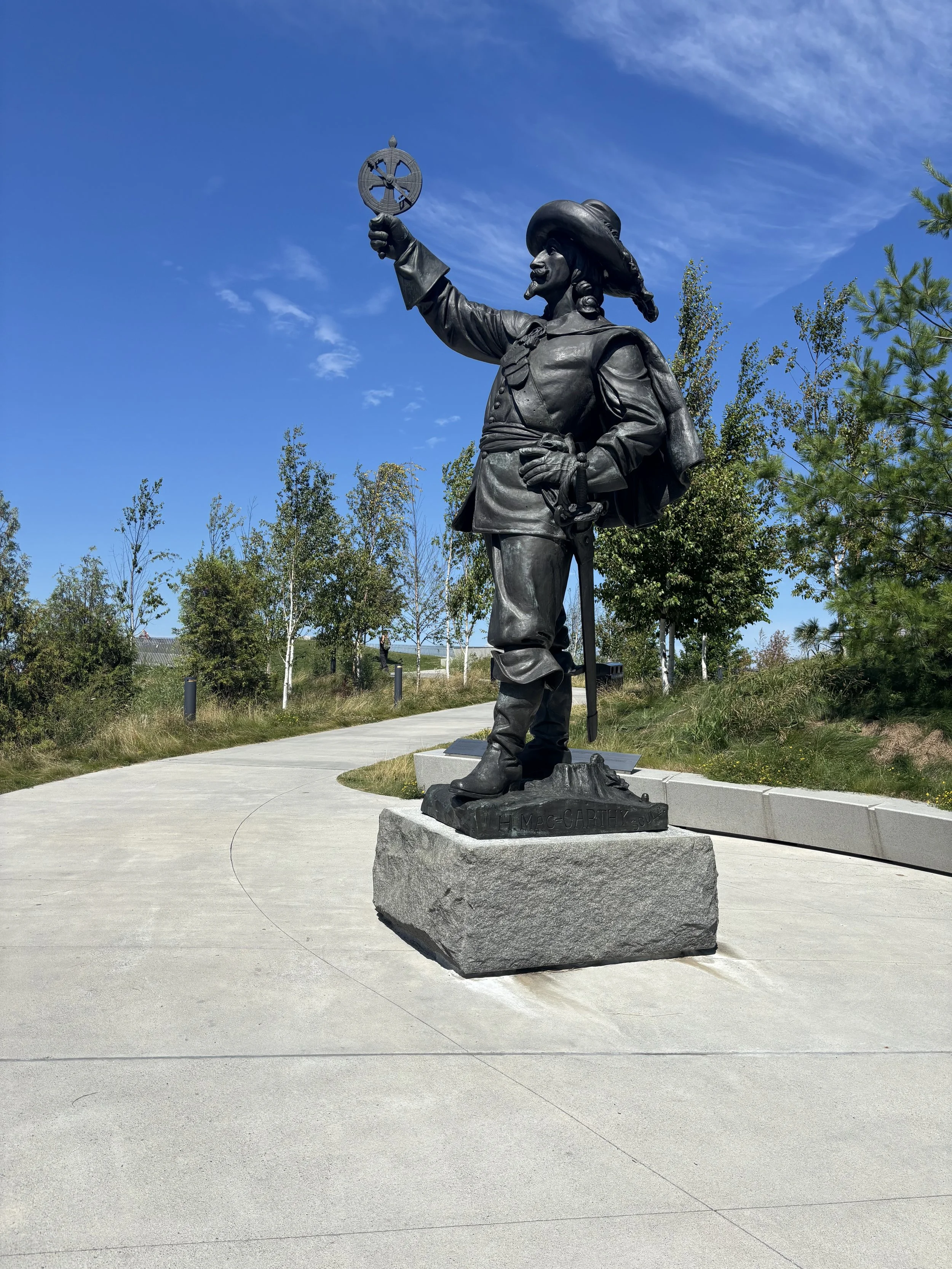 Bronze statue of a man in historic clothing, holding a shield with a cross symbol, standing outdoors on a stone pedestal, with a curved walkway and trees in the background under a blue sky.