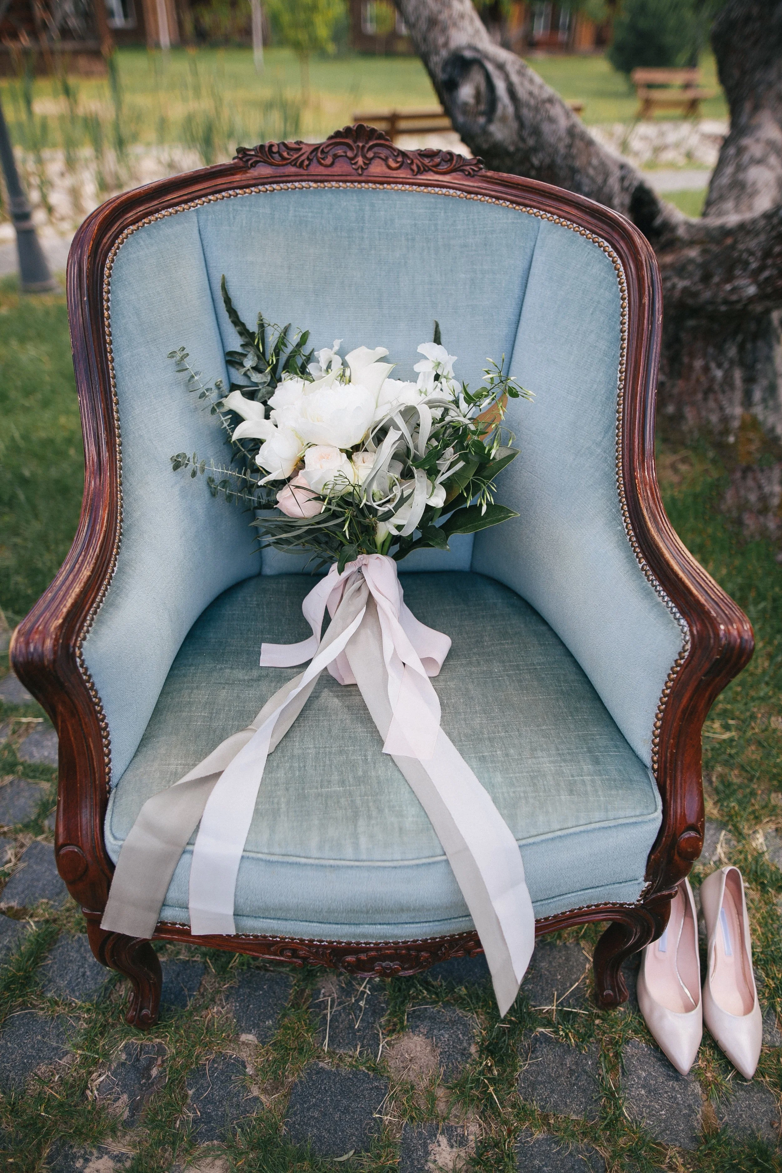 Un bouquet de fleurs blanches posé sur un fauteuil ancien en bois avec tissu bleu clair, à l'extérieur, avec des sandales beige à côté.