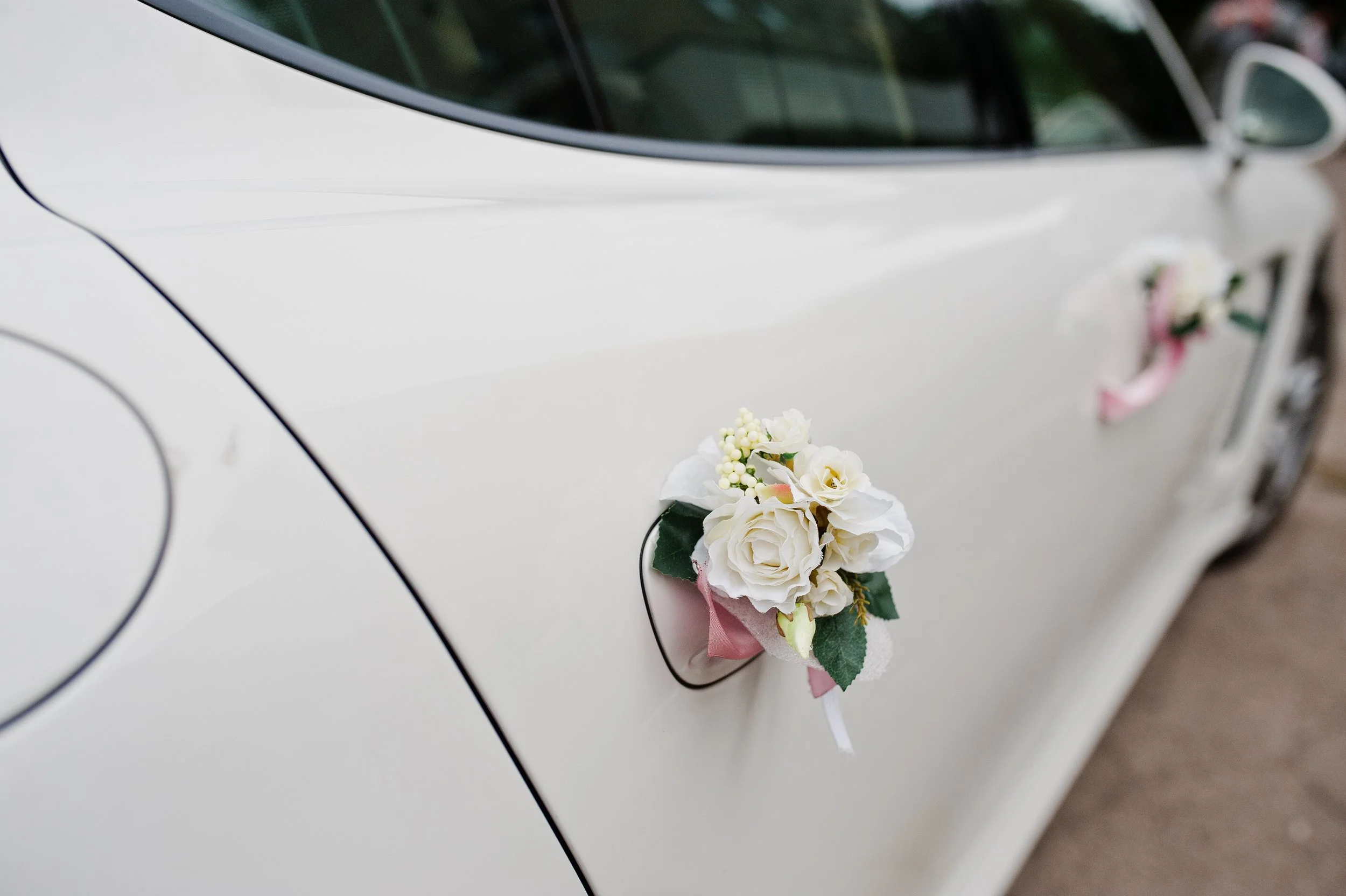 Voiture blanche décorée avec des bouquets de fleurs blanches et des rubans roses, probablement pour un mariage.