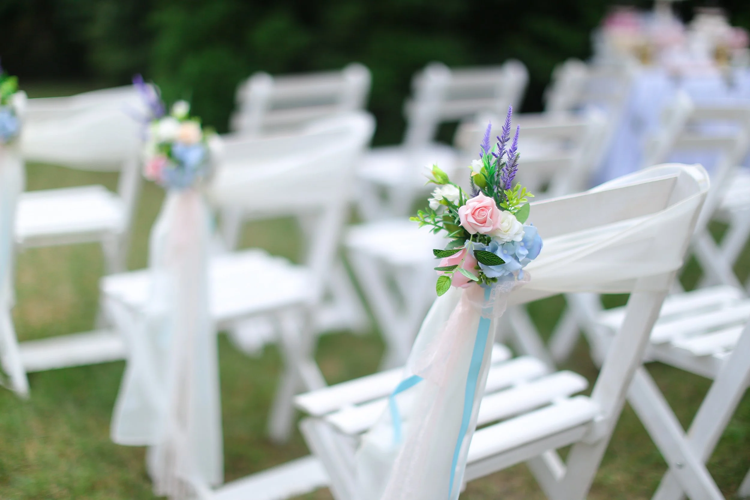 Chaises blanches décorées avec des bouquets de fleurs roses, blanches et violettes lors d'une cérémonie en plein air.