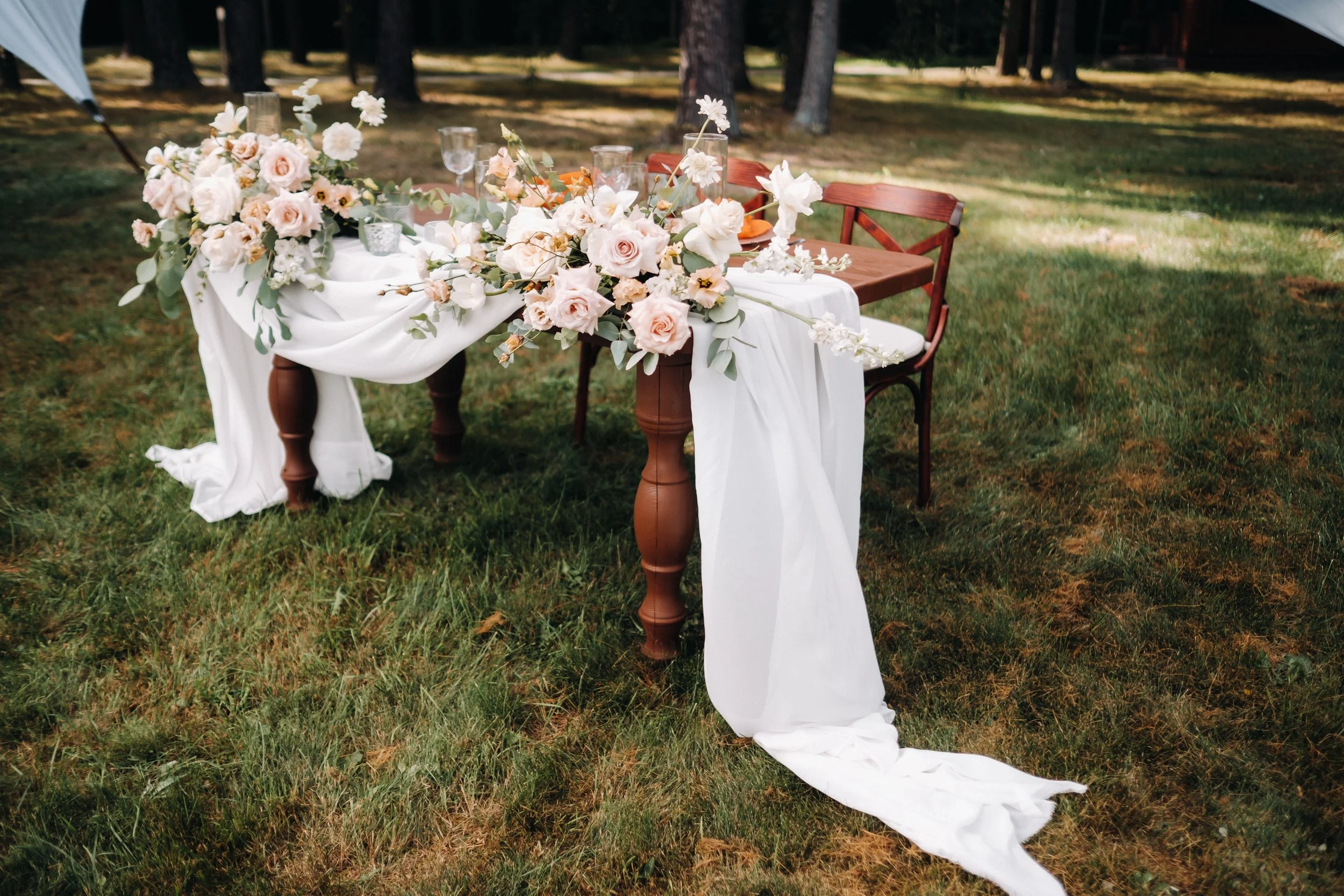 Table de mariage décorée avec un drapé blanc et un arrangement floral de roses et autres fleurs pâles, situé en plein air dans un espace vert avec des arbres en arrière-plan.