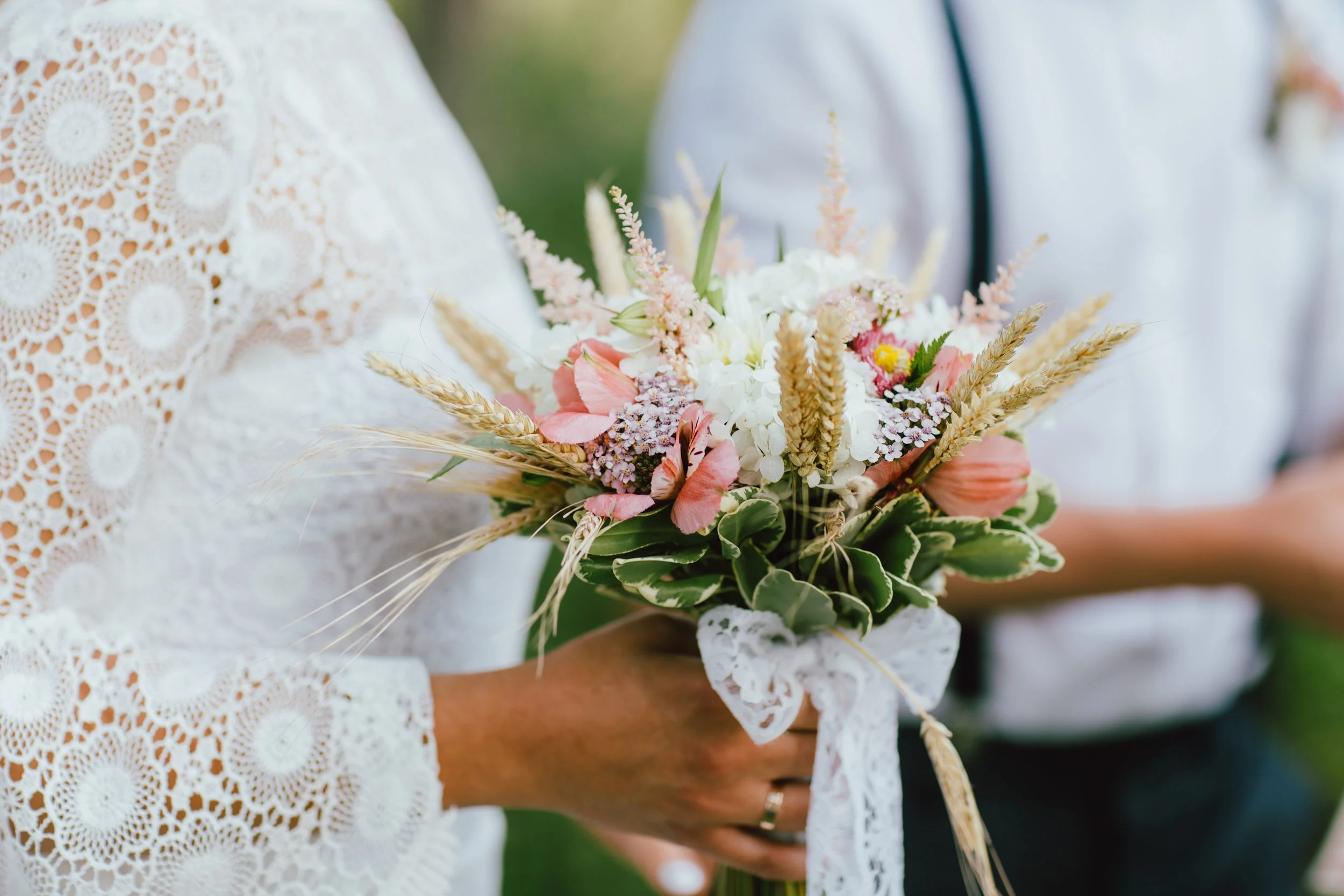 Une personne tenant un bouquet de fleurs avec des éléments champêtres, lors d'un mariage, la personne porte une robe en dentelle blanche, fond flou