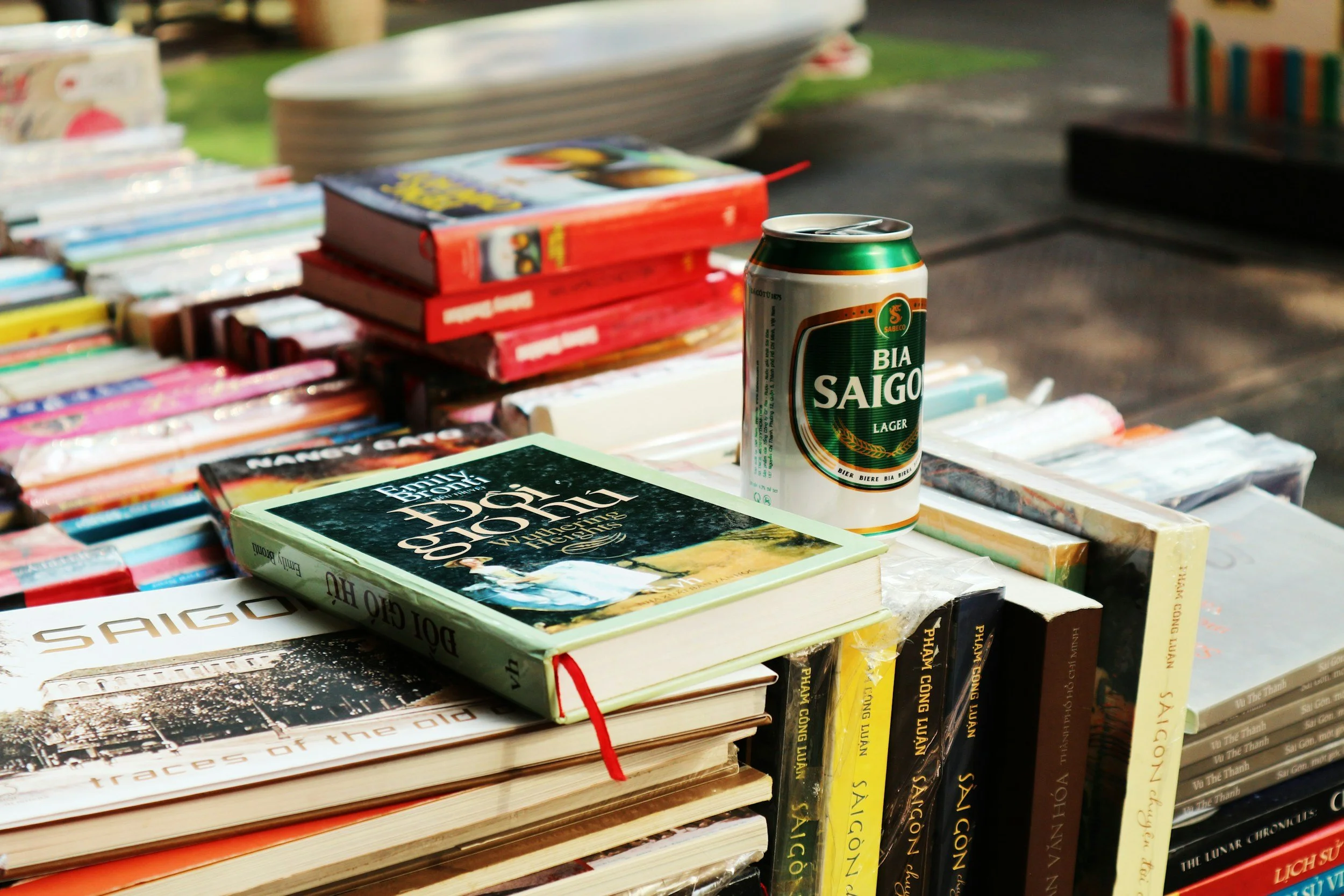 A can of Bia Saigon lager beer placed on top of a stack of books and magazines at an outdoor market or book sale.