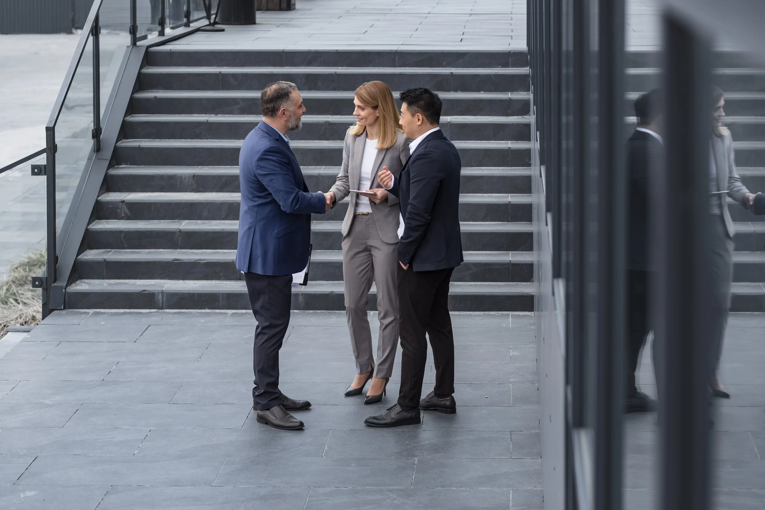 Three business professionals standing outside near a staircase, shaking hands and engaging in conversation.