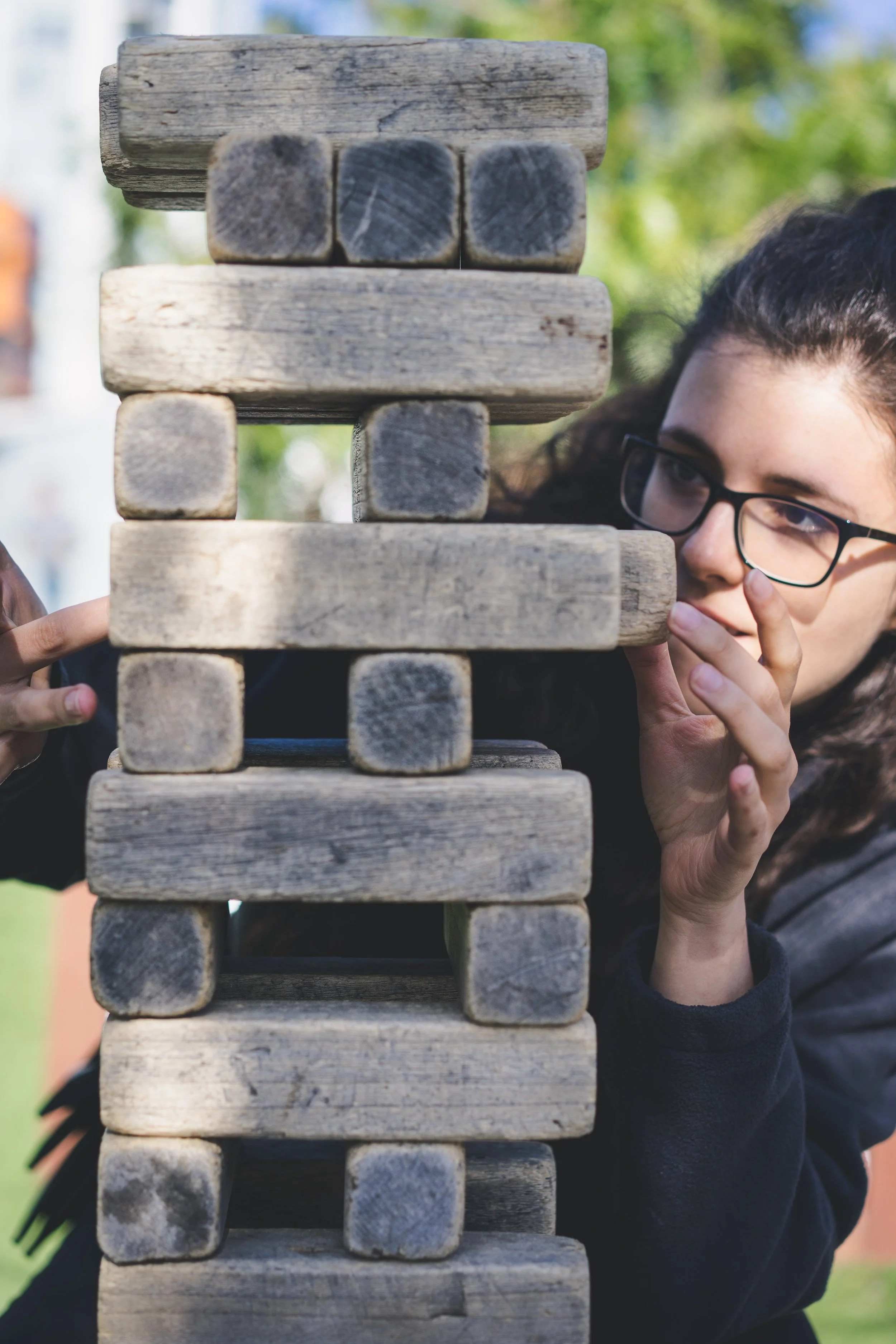 A woman with glasses playing outdoor Jenga, carefully removing a wooden block from the tower.