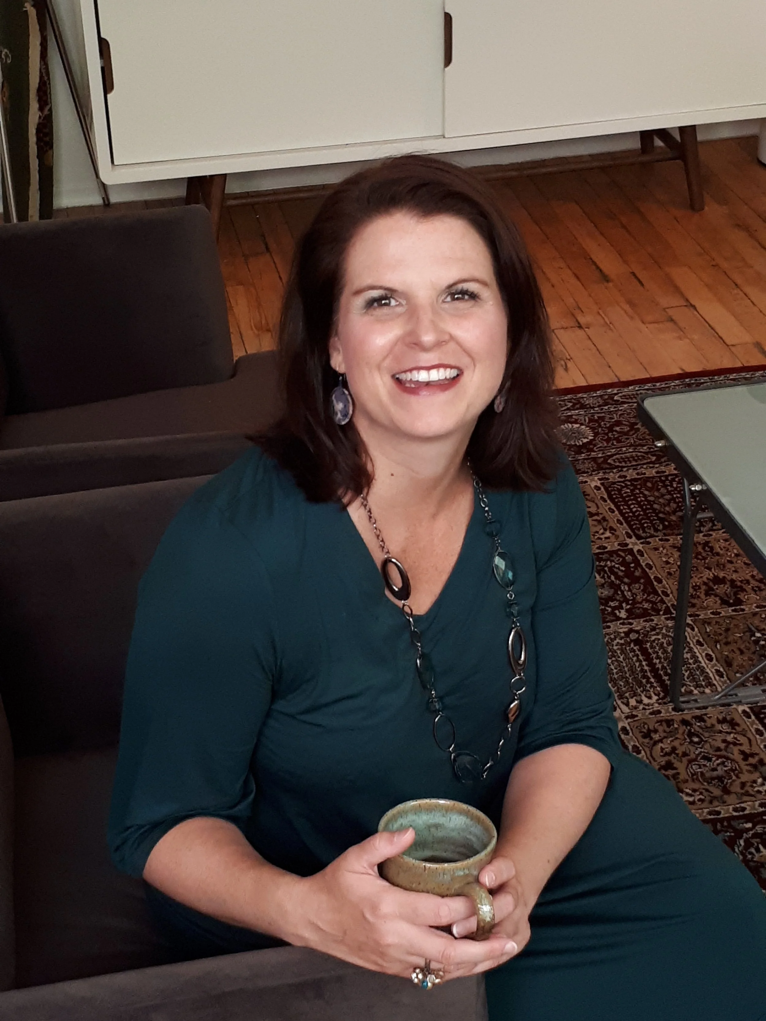 A woman with dark brown hair is sitting on a brown couch, smiling and holding a ceramic mug, wearing a teal dress and jewelry, in a room with wooden floors and a white cabinet in the background.