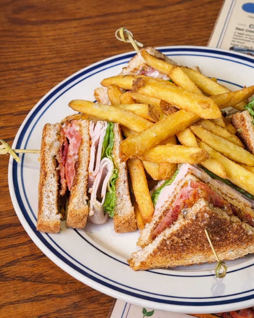 A plate with fries and a club sandwich cut into quarters. The sandwich contains turkey, ham, bacon, lettuce, tomato, and mayonnaise on toasted bread. The plate is on a wooden table.