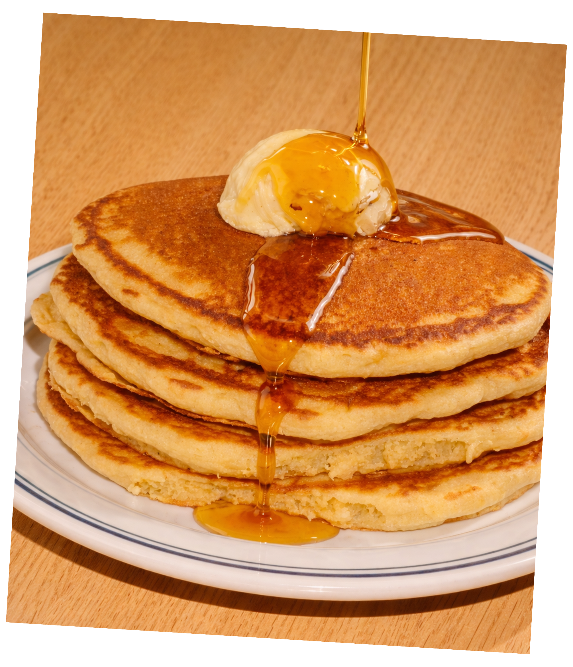 Stack of four thick pancakes topped with a scoop of butter, drizzled with syrup, with syrup being poured over.