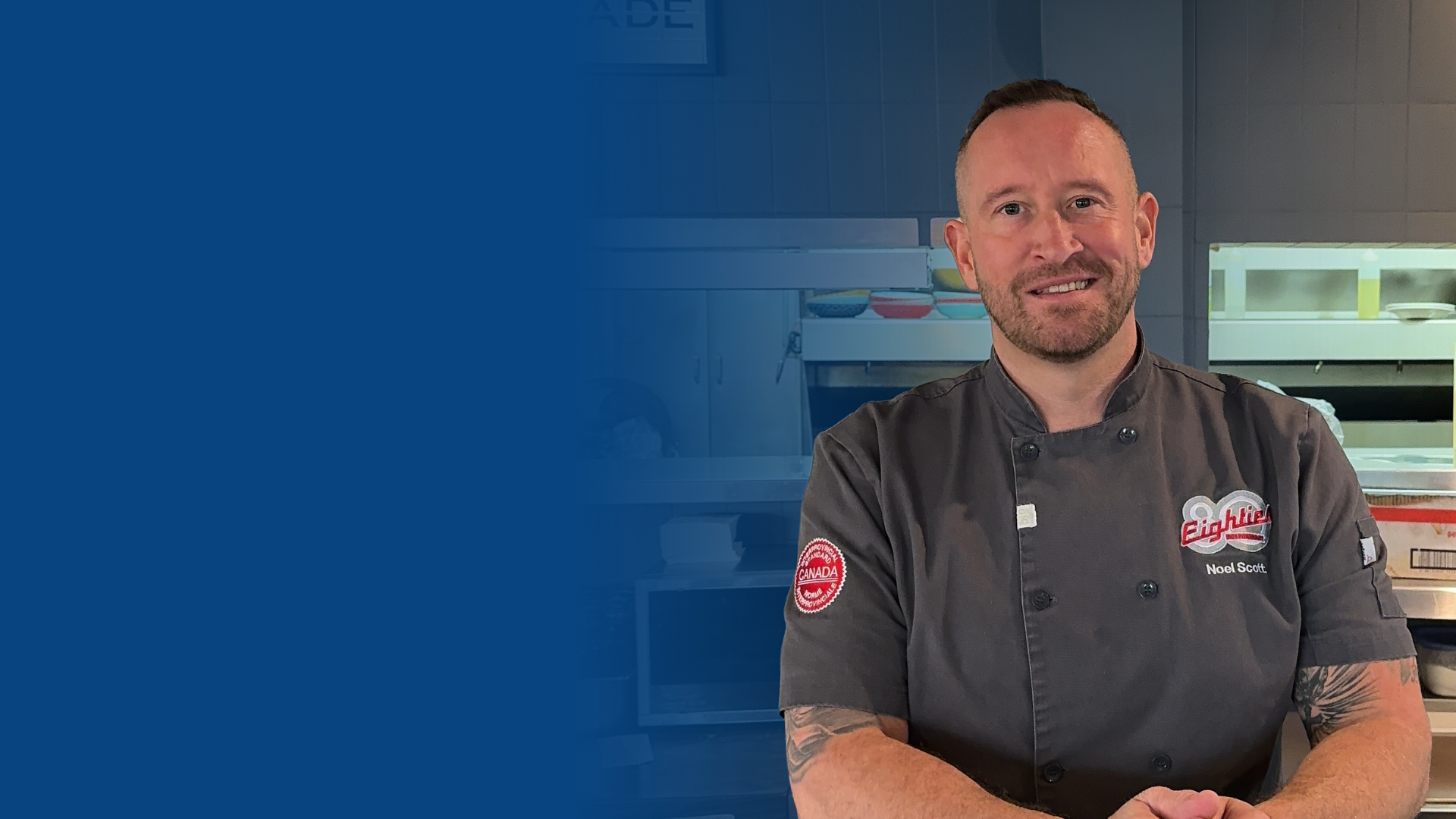 A male chef in a professional kitchen, wearing a black chef's coat with patches and the name 'Noel Scott' embroidered on it, smiling with arms crossed.