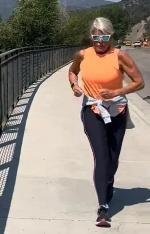 An elderly woman wearing sunglasses and an orange sleeveless top is jogging on a paved path beside a guardrail, with mountains and trees in the background.