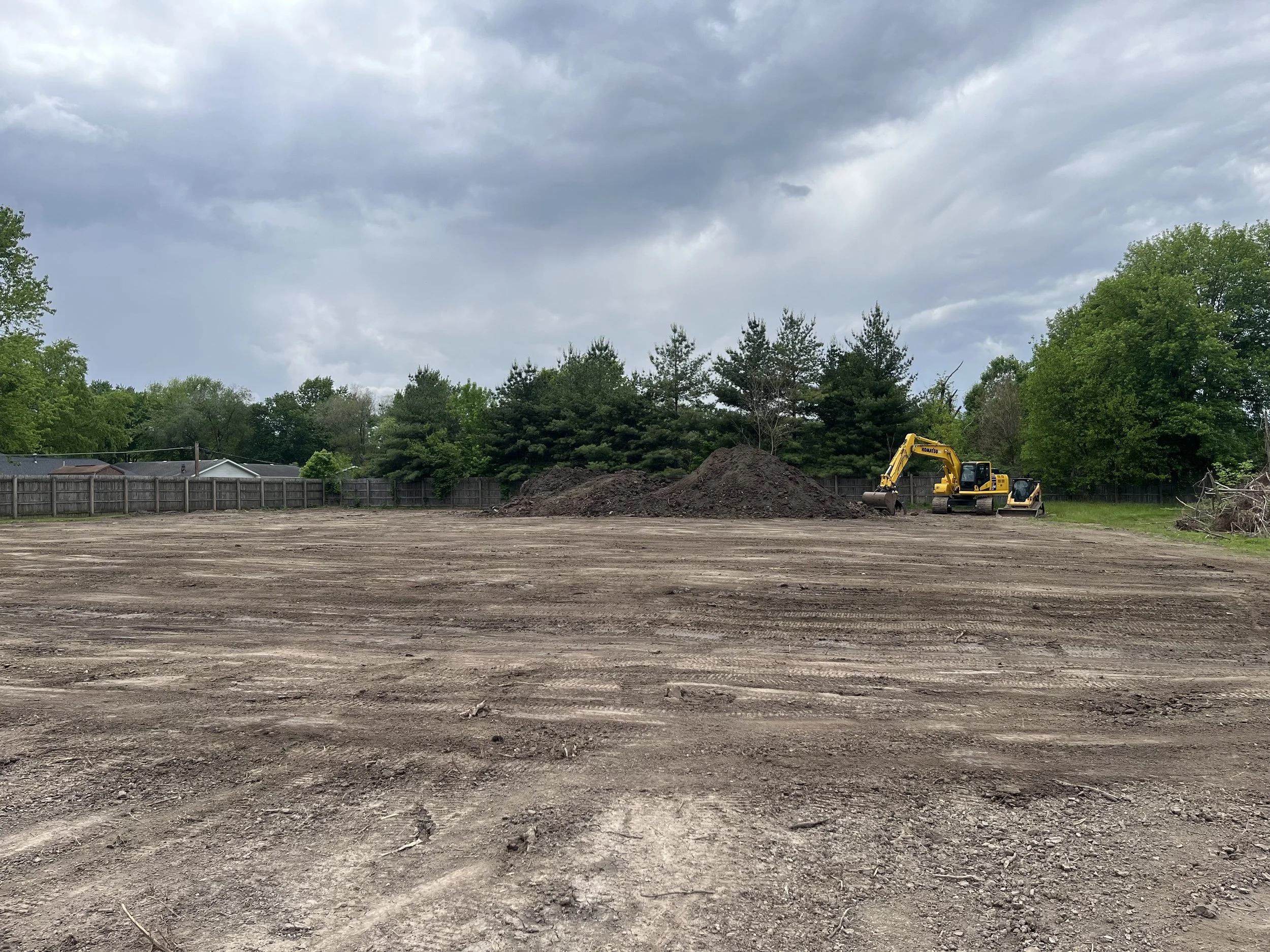 Construction site with excavator and piles of dirt, surrounded by trees and a cloudy sky.