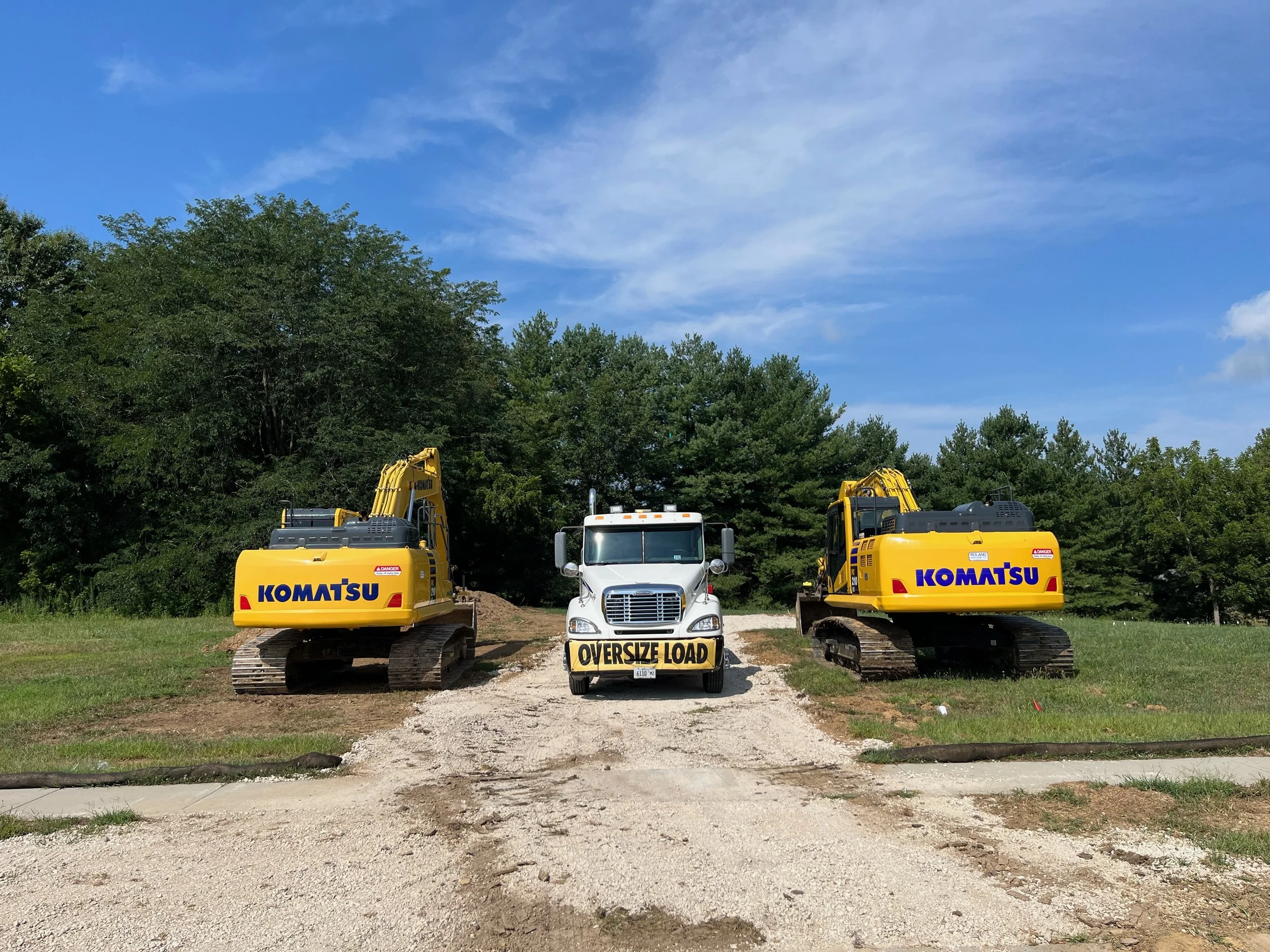 A construction site with a white truck labeled 'OverSize Load' between two yellow Komatsu excavators on a gravel path, with a background of trees and a blue sky with some clouds.