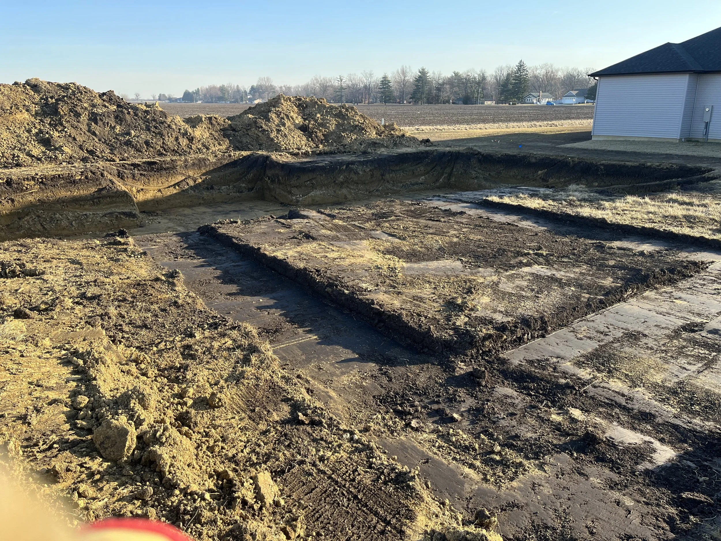 A construction site with excavated dirt and a cleared area in a backyard, with a house, trees, and fields in the background during daytime.
