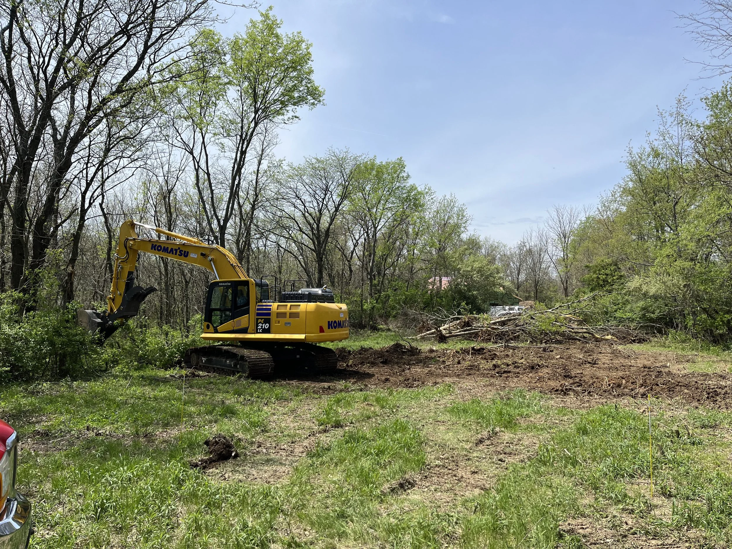 A yellow Komatsu excavator is clearing a wooded area, with trees and a partly cloudy sky in the background.